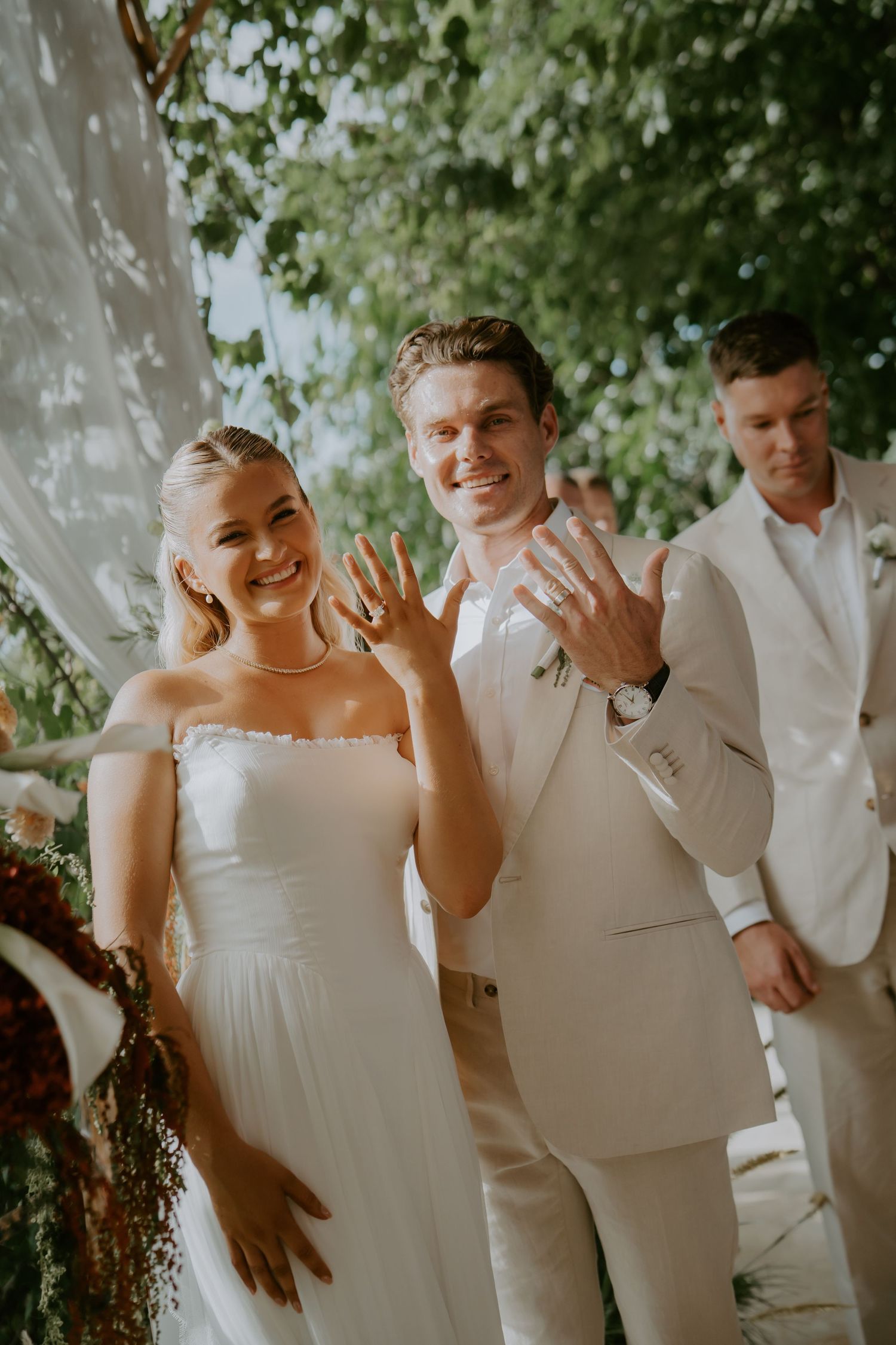 Newlyweds joyfully display wedding rings at outdoor tropical ceremony celebration.