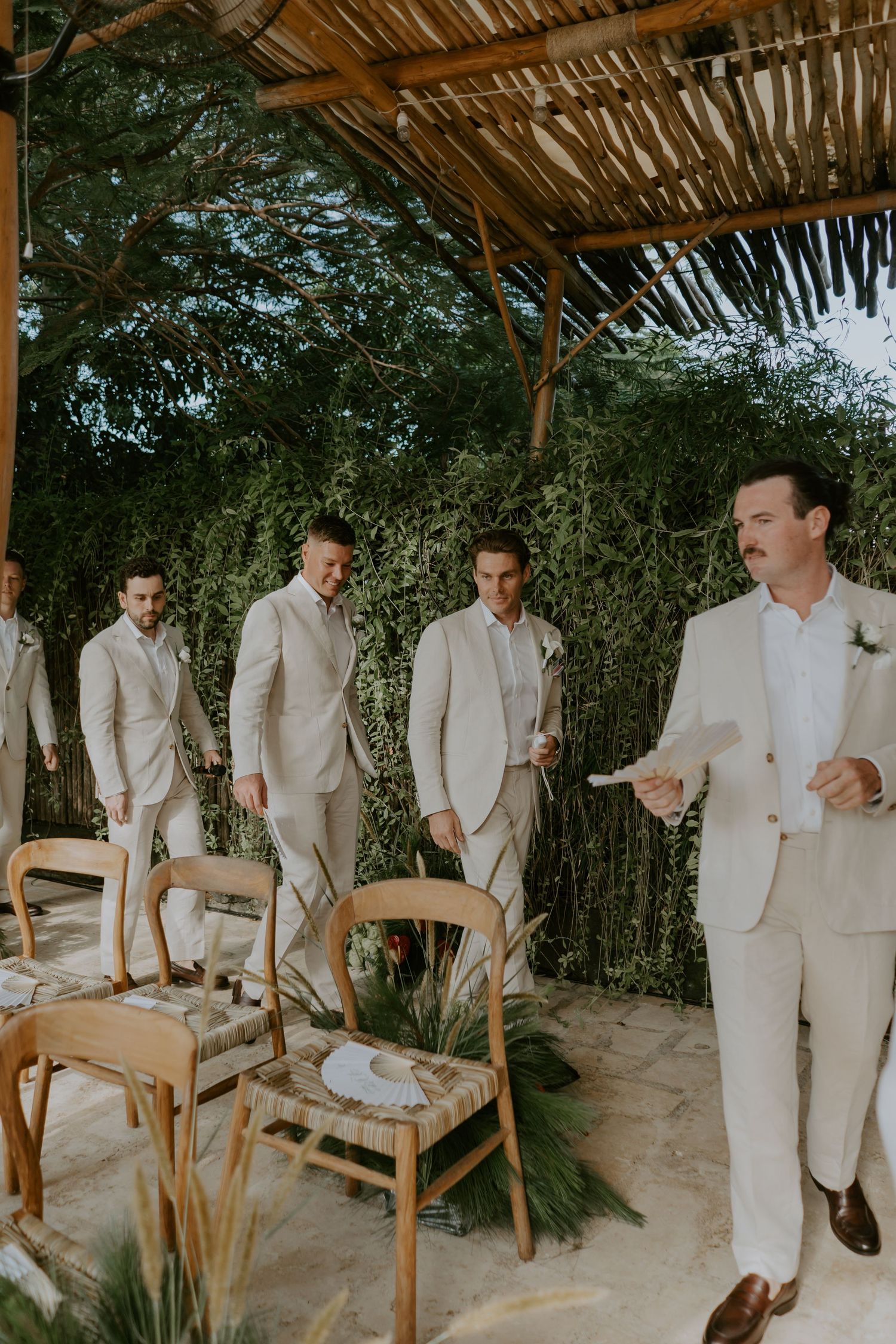 Groomsmen in beige suits gather under bamboo pergola with lush greenery backdrop at outdoor wedding ceremony.