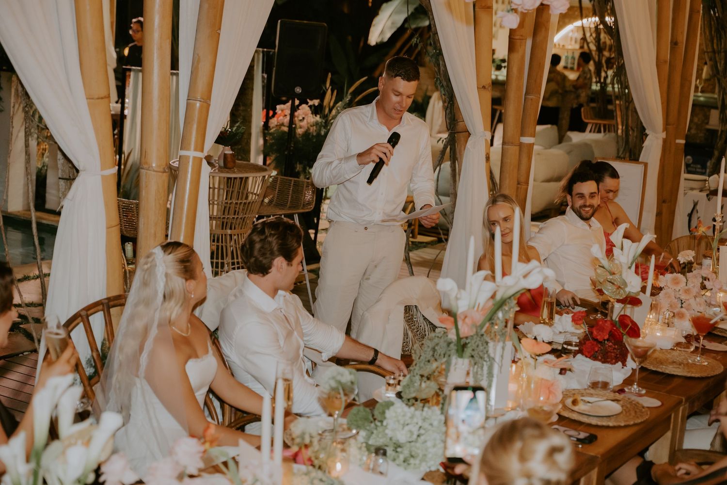Guest delivers speech at elegant outdoor wedding reception while bride and groom listen smiling.