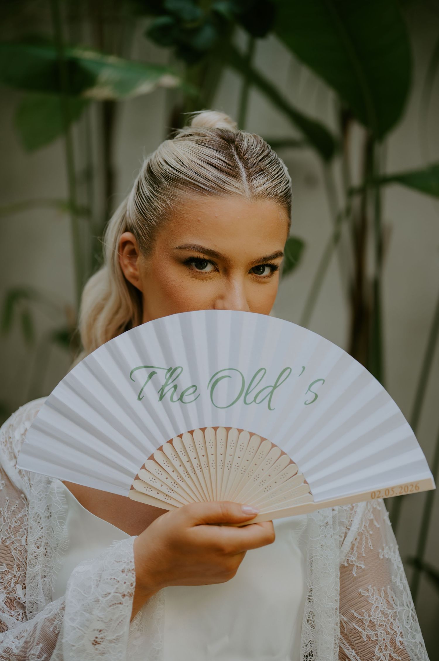 Blonde bride holding white personalized fan reading The Olds in green script before tropical greenery.