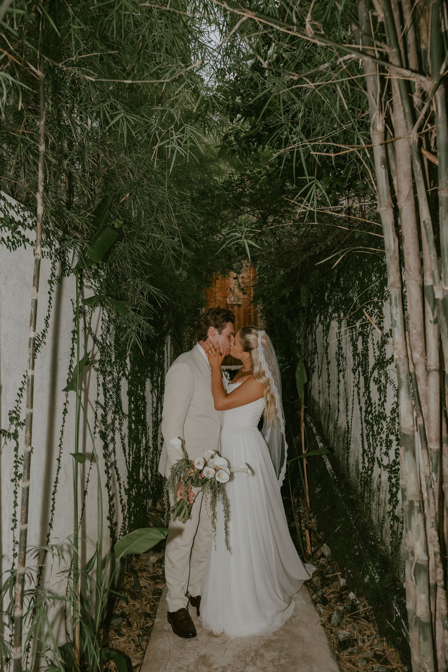 Bride and groom share romantic kiss in lush bamboo garden pathway at night.