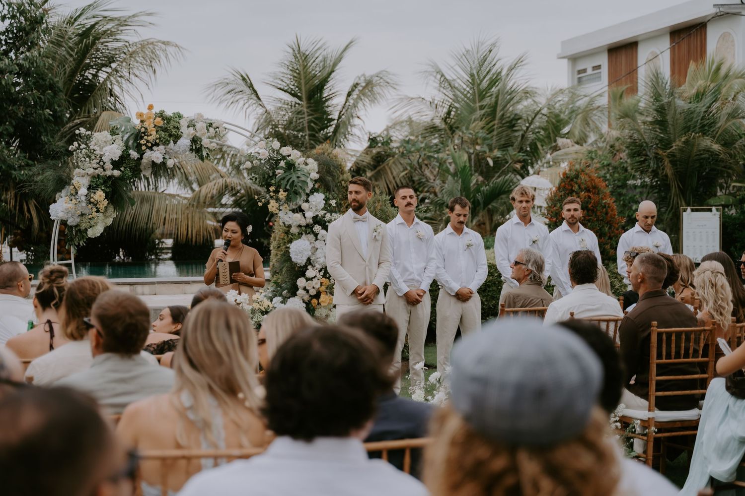 Groom and groomsmen in white suits stand at outdoor wedding altar surrounded by palm trees.