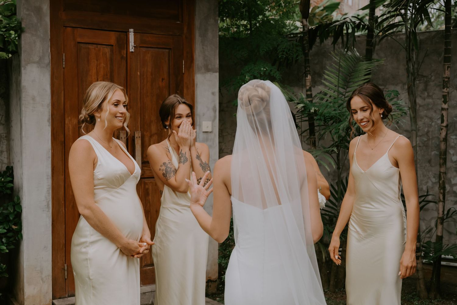 Bride in white gown and veil shares emotional first look moment with three bridesmaids in ivory dresses.