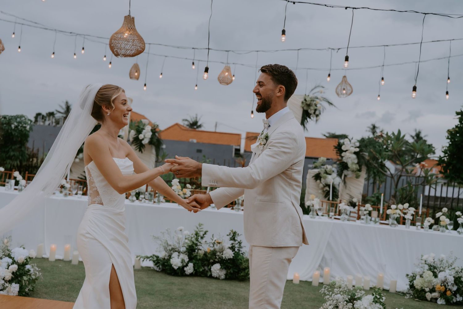 Bride and groom hold hands and dance at outdoor reception under string lights at dusk.
