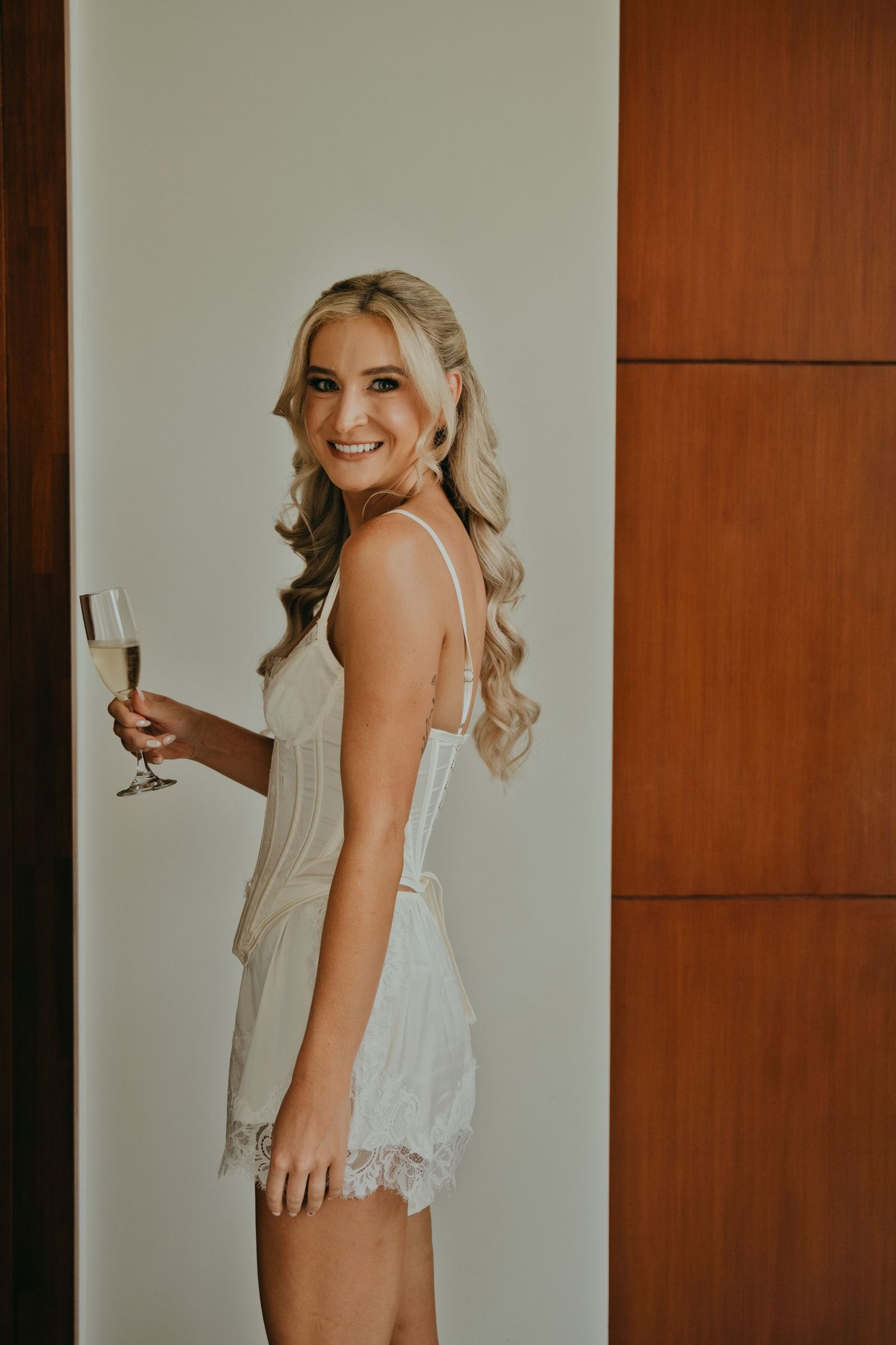 Smiling blonde bride in white lace romper holding a champagne glass during wedding morning preparations.