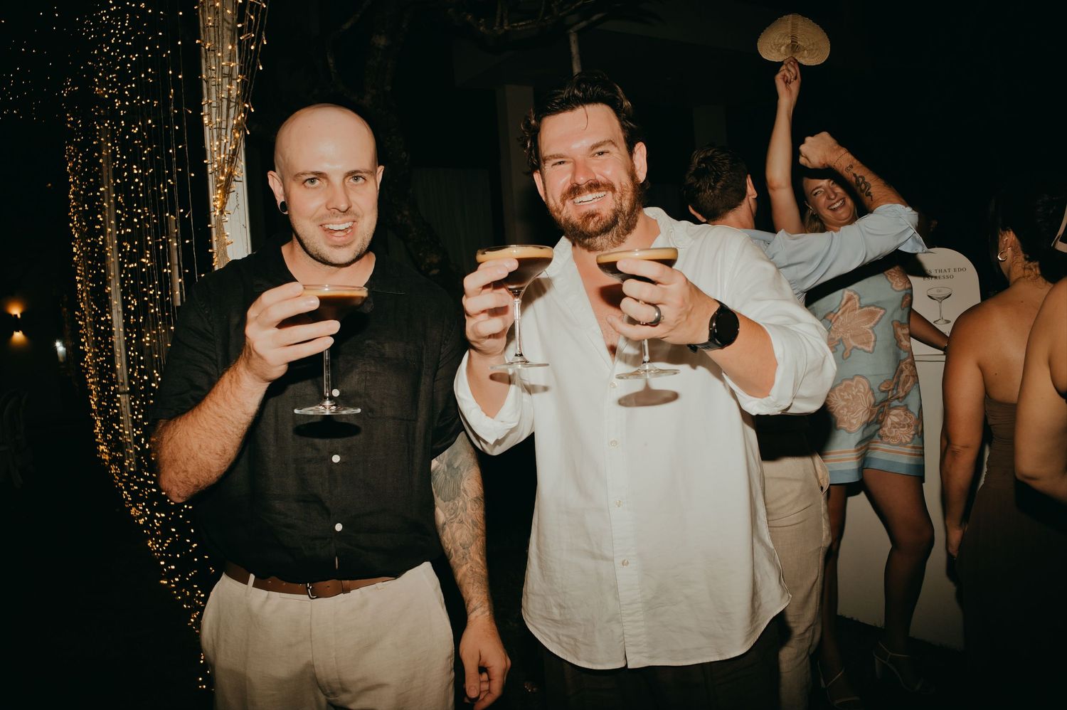 Two male wedding guests smile and raise cocktail glasses in a toast at evening reception.