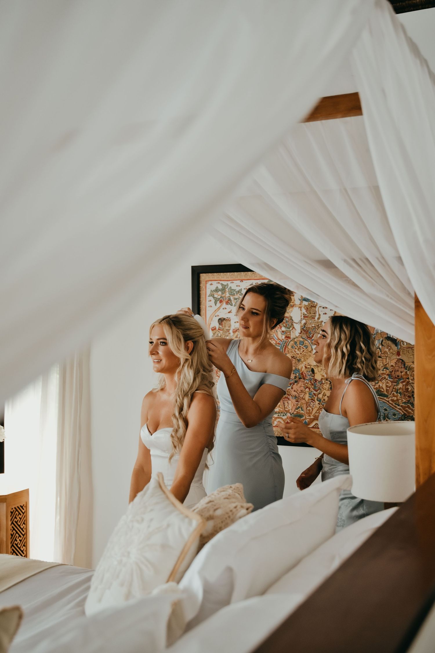Bridesmaids in blue dresses helping blonde bride with hair under white canopy bed before wedding.