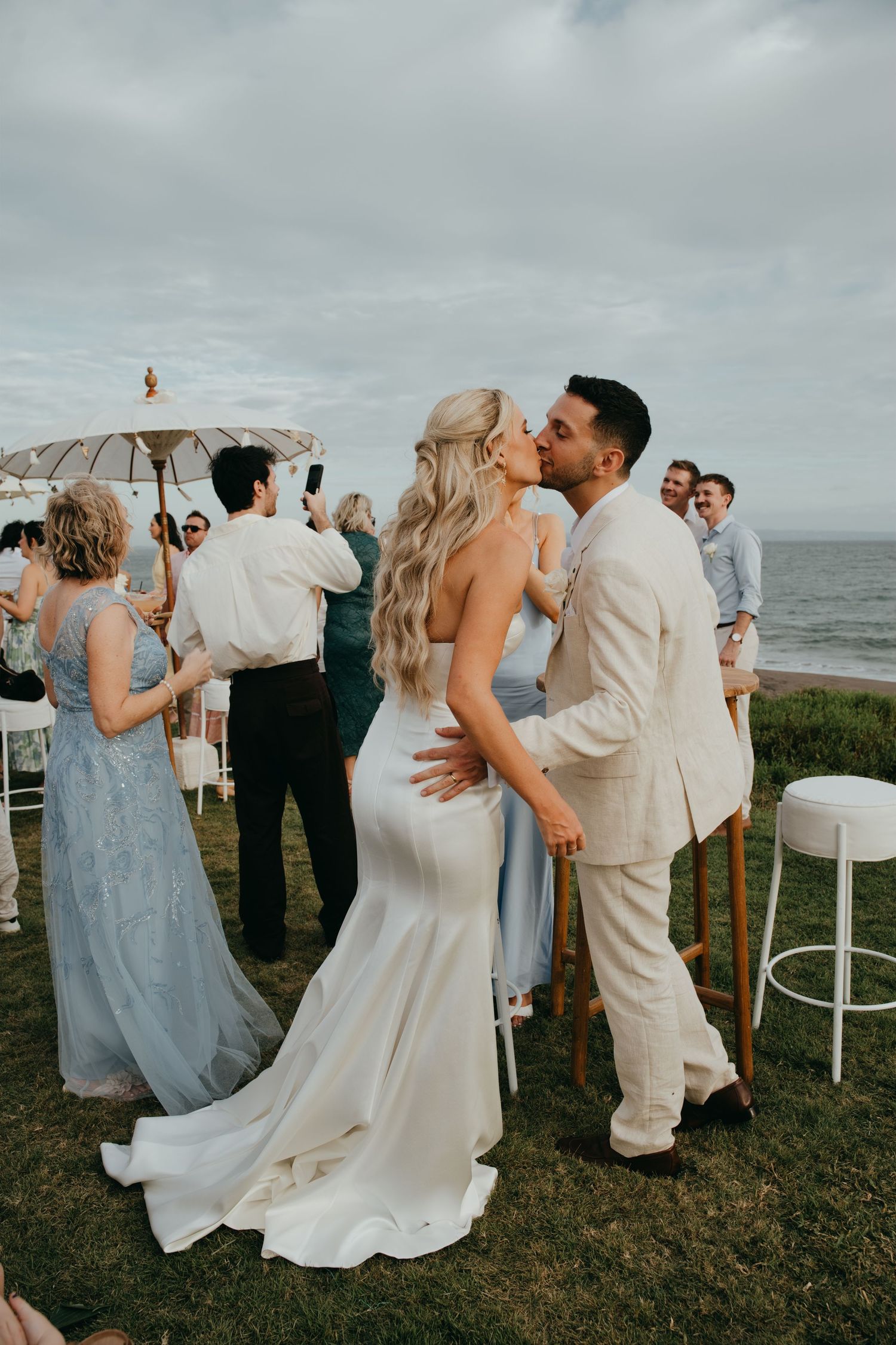 Newlyweds sharing a kiss surrounded by guests at outdoor oceanside wedding reception celebration.