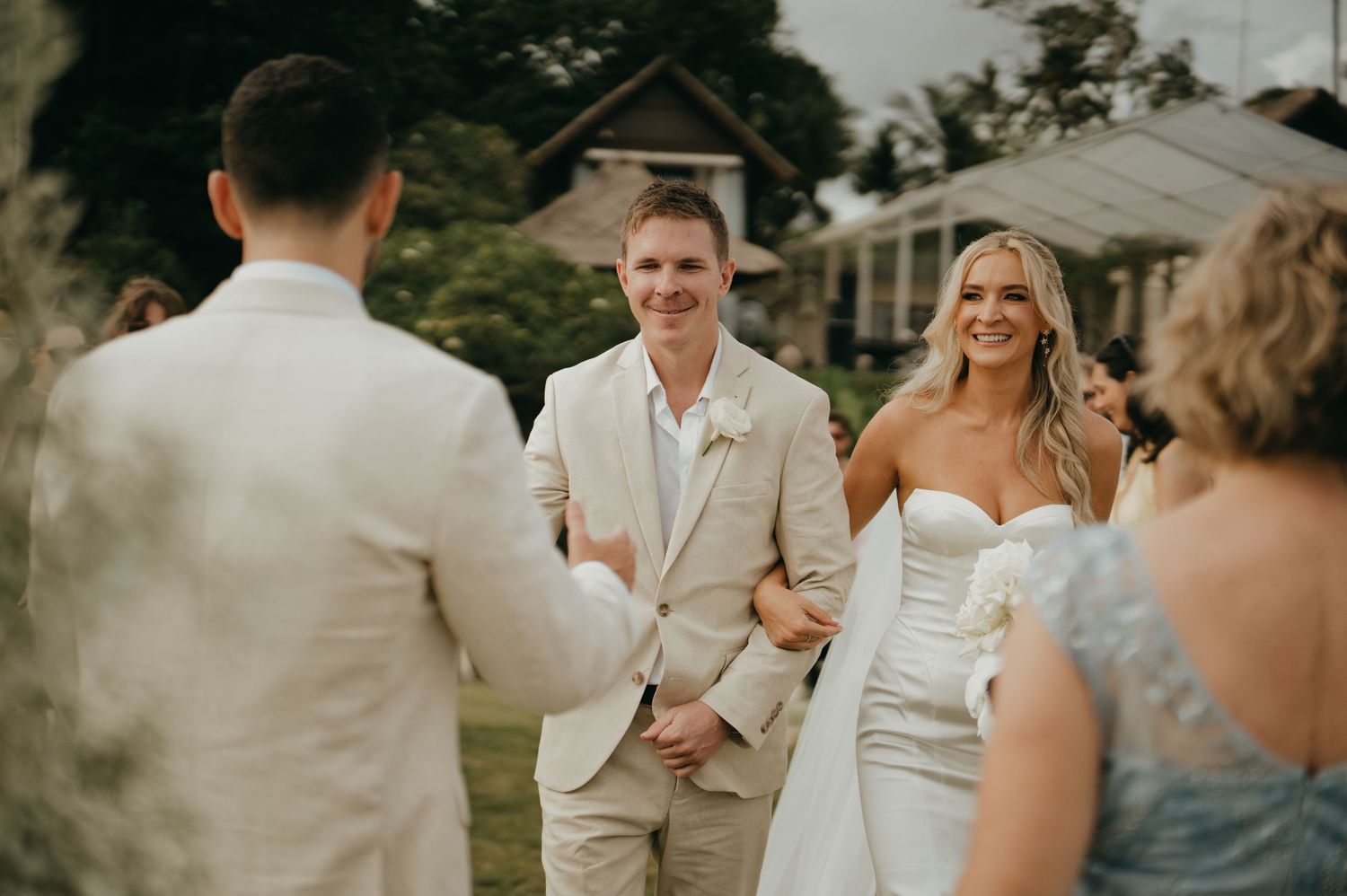 Smiling bride in strapless white gown walks toward groom at outdoor wedding ceremony.