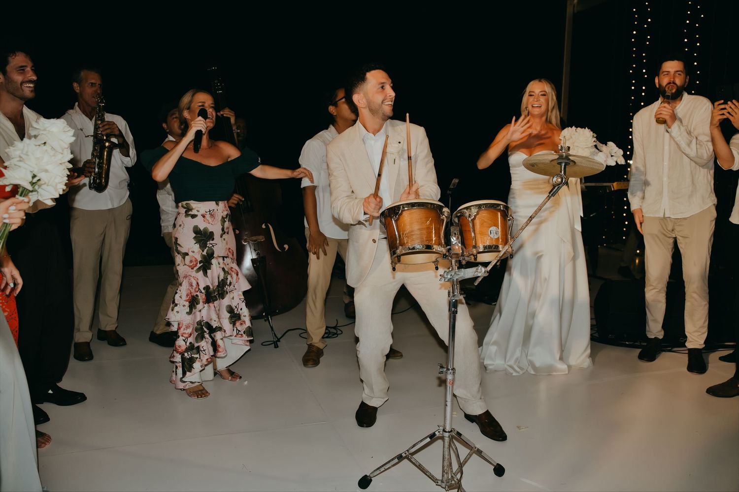 Groom plays drums energetically on the dance floor surrounded by celebrating wedding guests at night.
