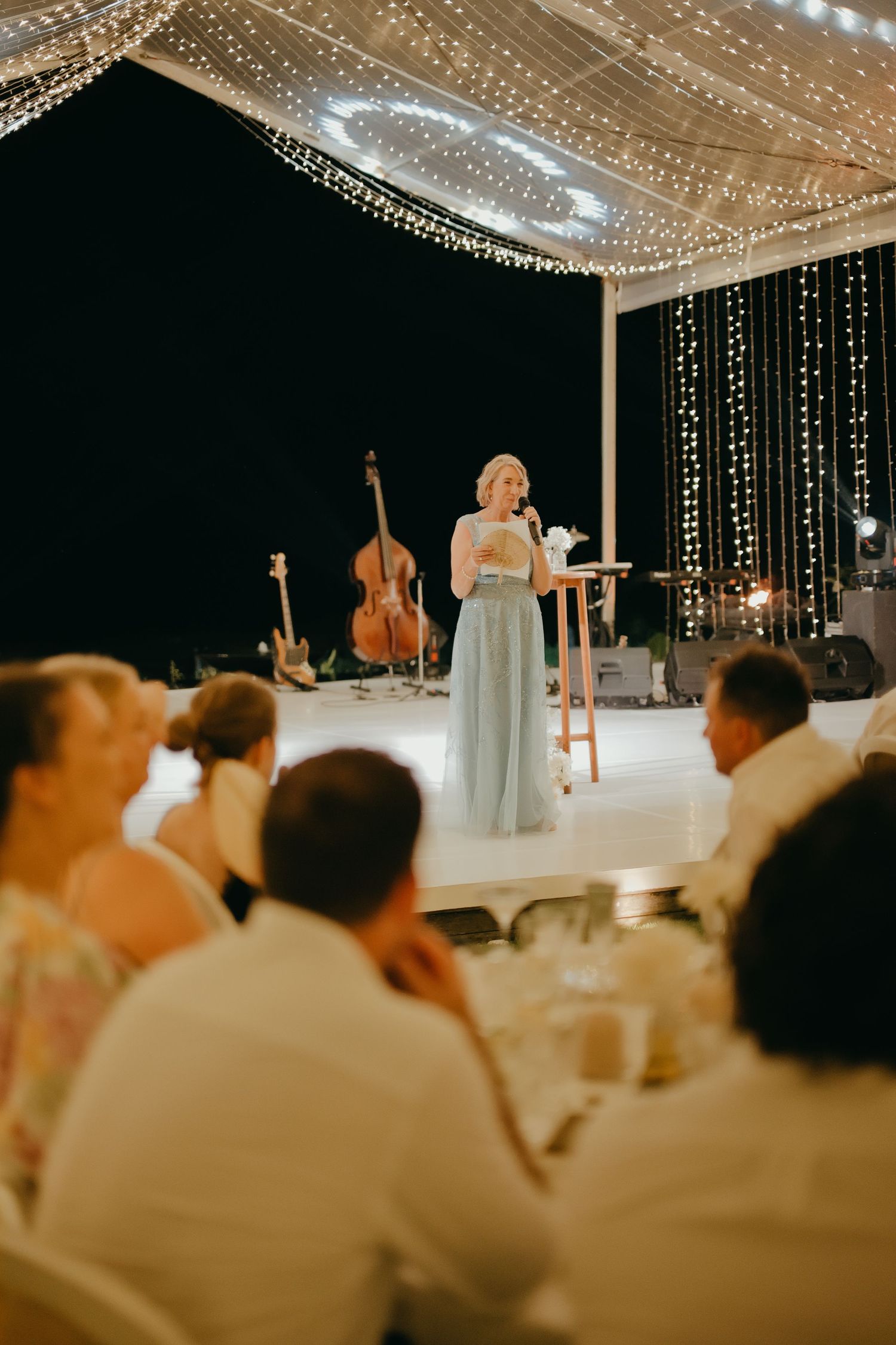 Wedding guest in blue gown delivers a heartfelt speech on a fairy-light-adorned stage at night.