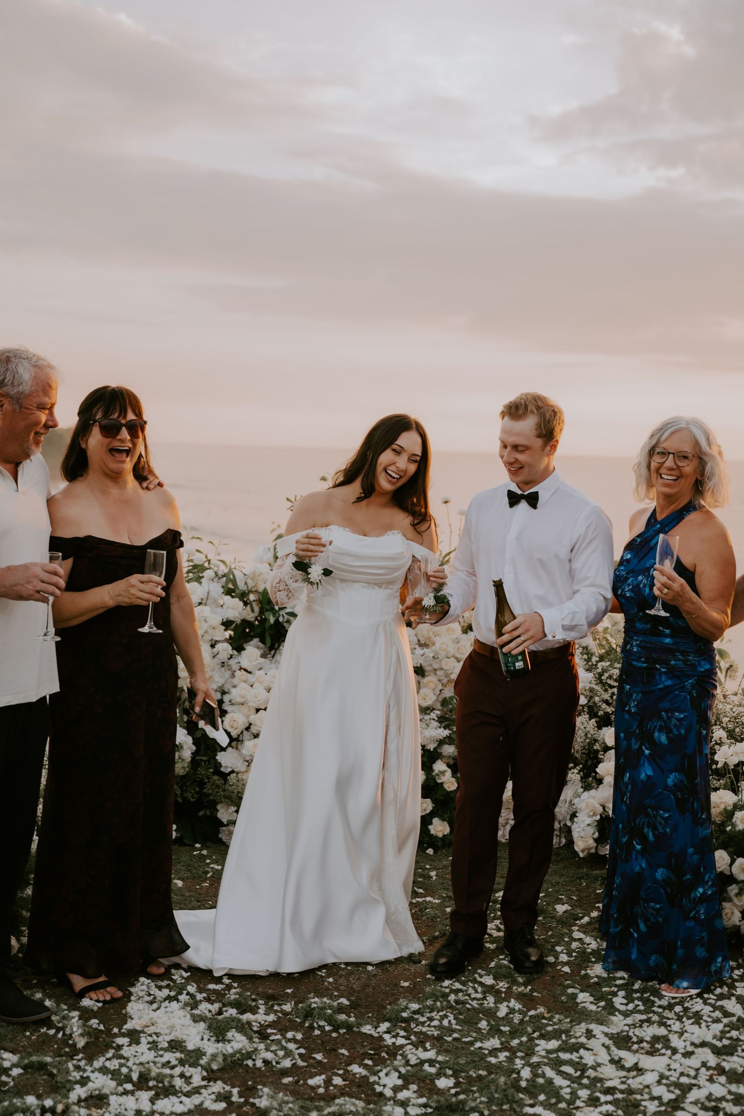 Bride and groom celebrate with family members holding champagne glasses during an outdoor wedding reception at sunset by the ocean.
