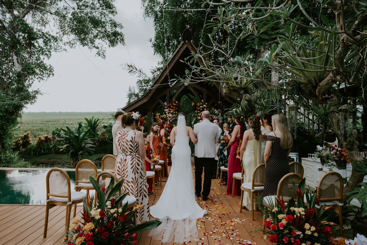 Bride walks down outdoor aisle surrounded by guests at a tropical garden wedding ceremony.