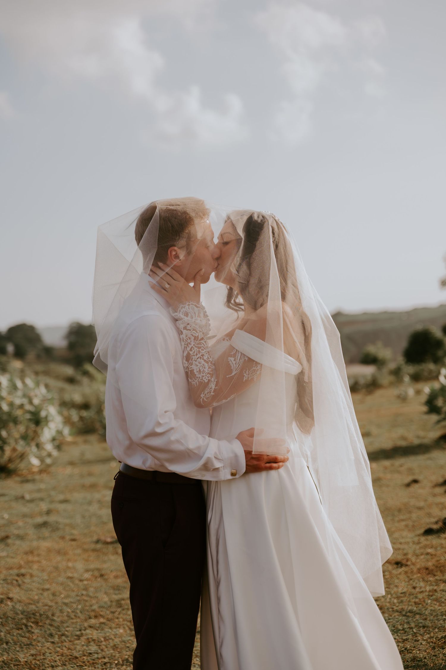 Newlywed couple sharing romantic kiss under bride's veil at outdoor wedding with scenic countryside backdrop.