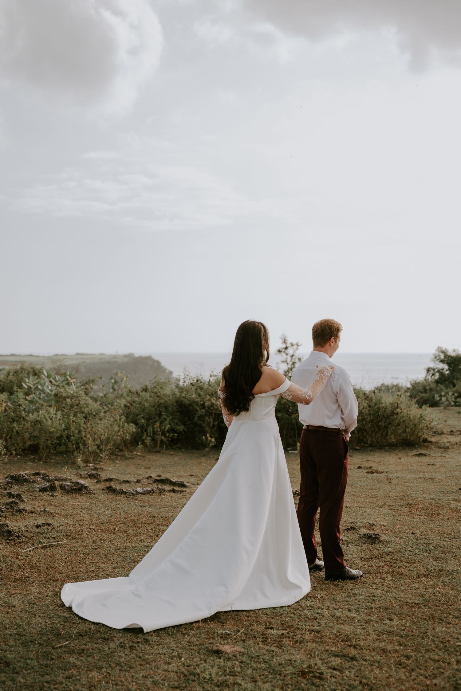 Bride in white off-shoulder gown and groom stand together overlooking ocean at cliffside wedding venue.