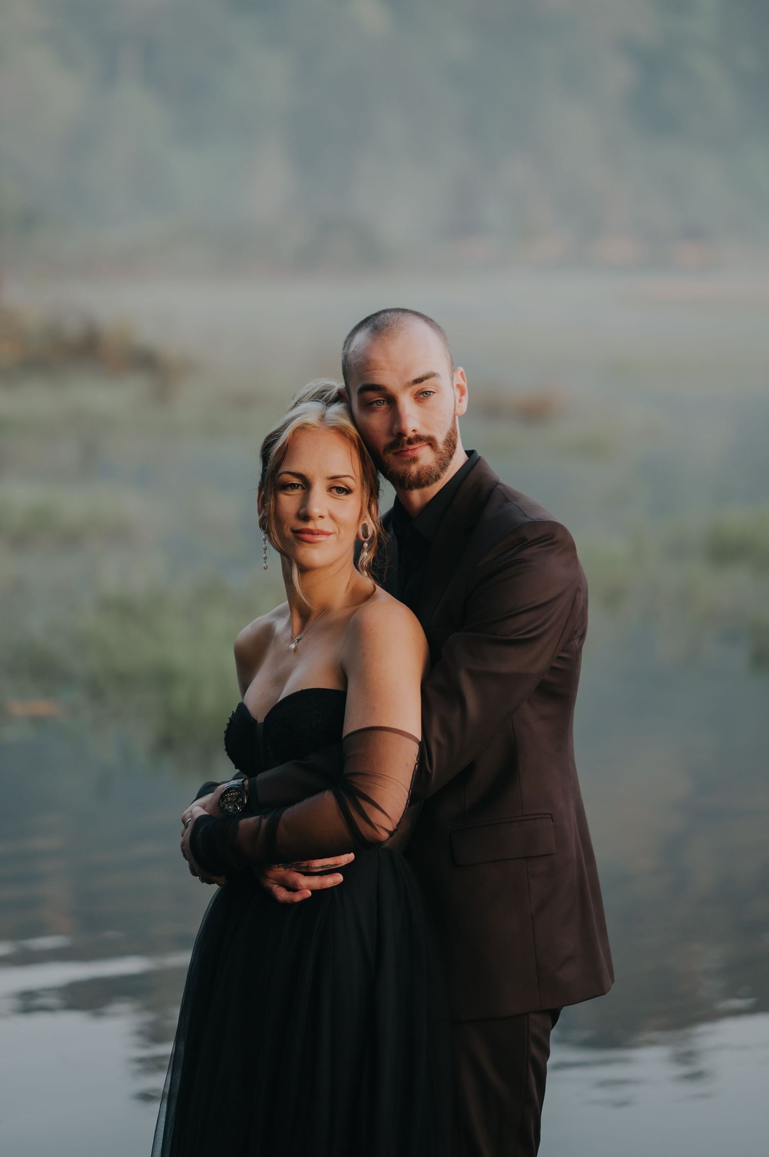 A romantic couple embraces in front of a misty lake wearing formal attire during a moody photoshoot.