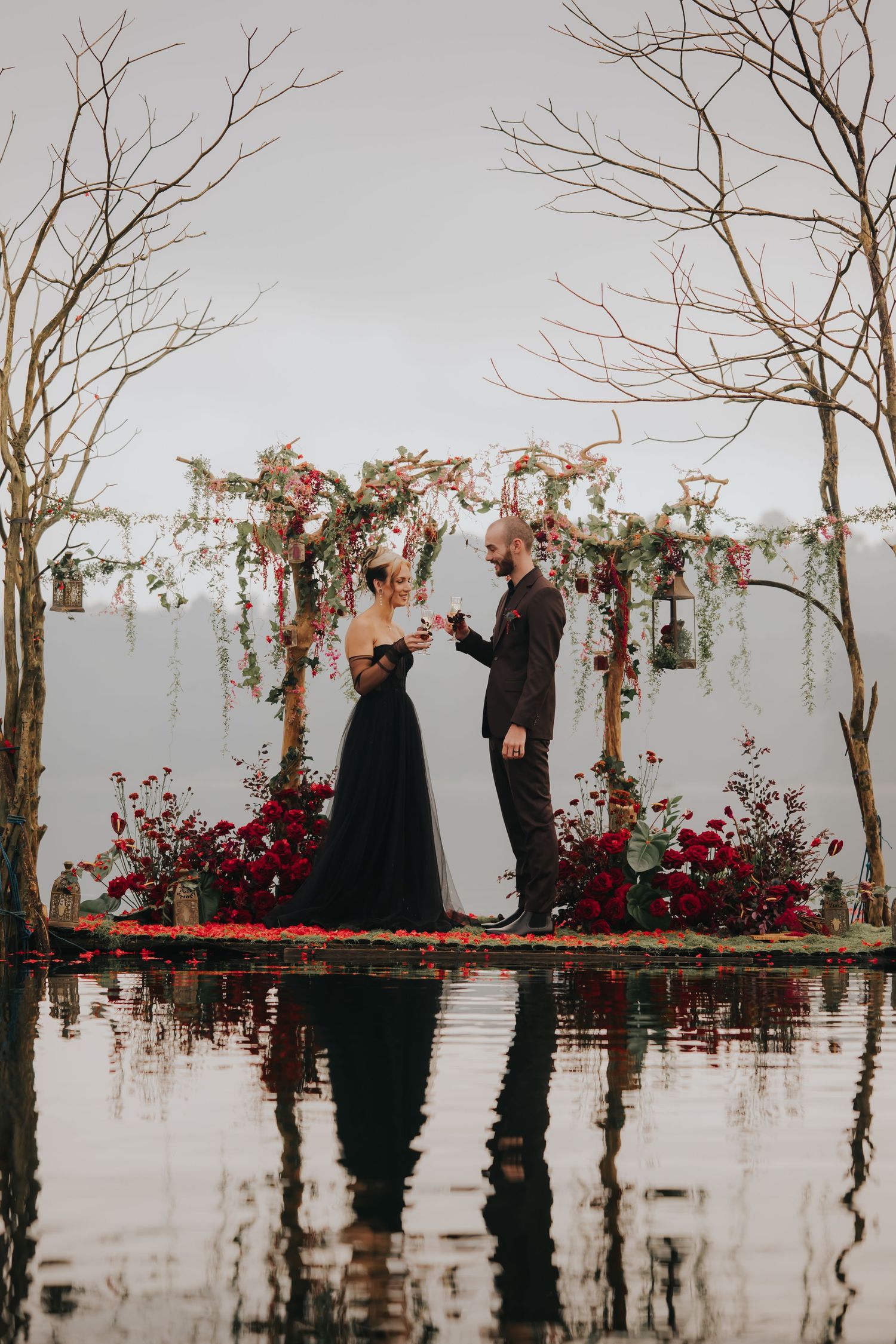 Misty lakeside wedding ceremony with bare trees and red flowers reflecting in dark waters.