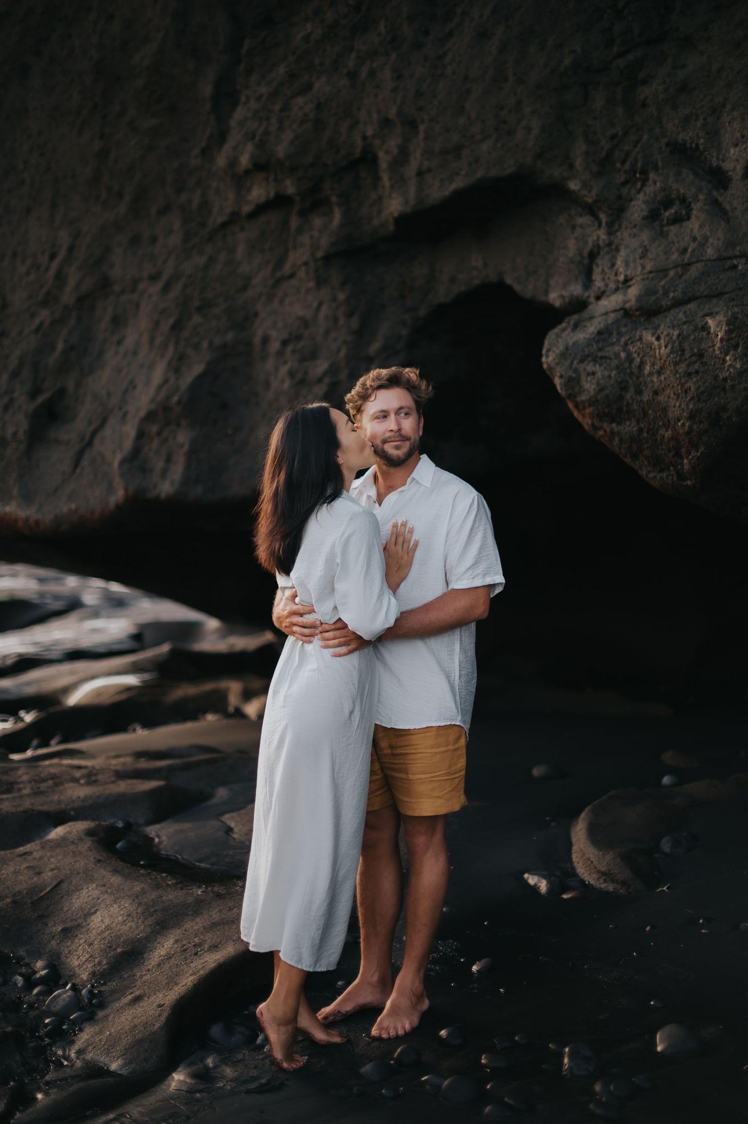 A couple embraces on dark rocky beach terrain at sunset in matching white outfits creating intimate silhouettes.