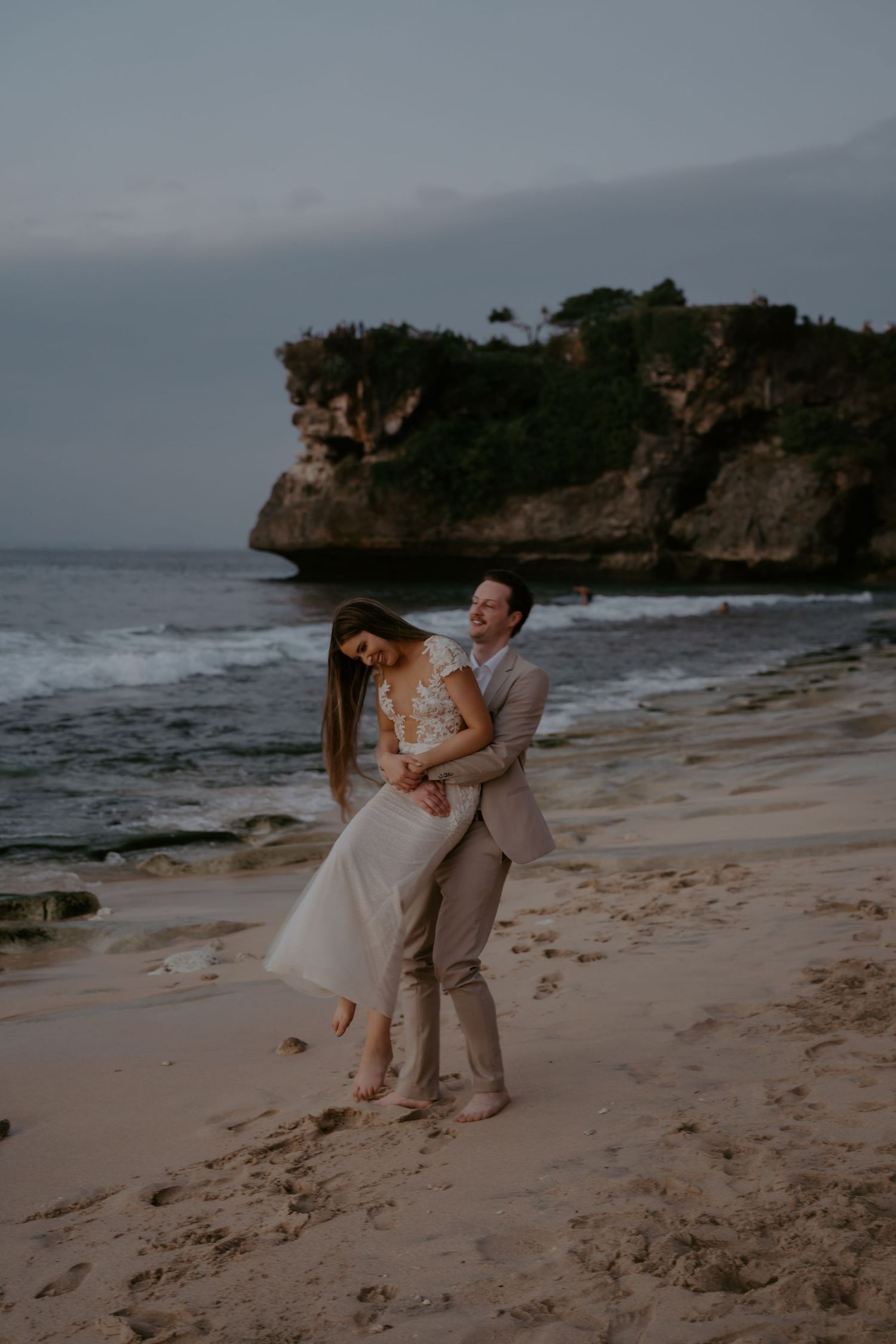 A couple in wedding attire shares romantic moments on a scenic beach with rocky cliffs in the background at sunset.