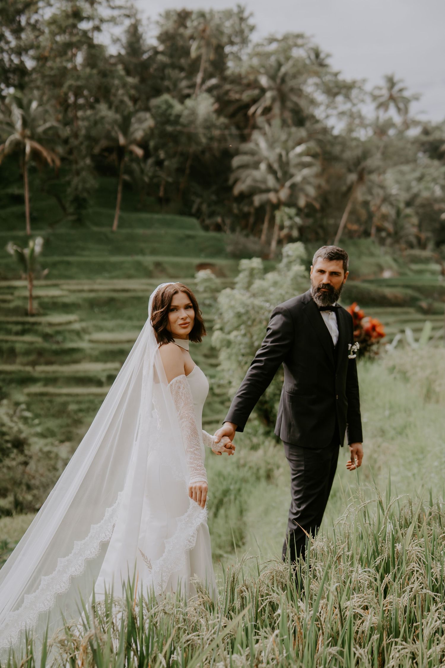 A bride and groom share an intimate moment in a lush green field surrounded by palm trees and tropical foliage.