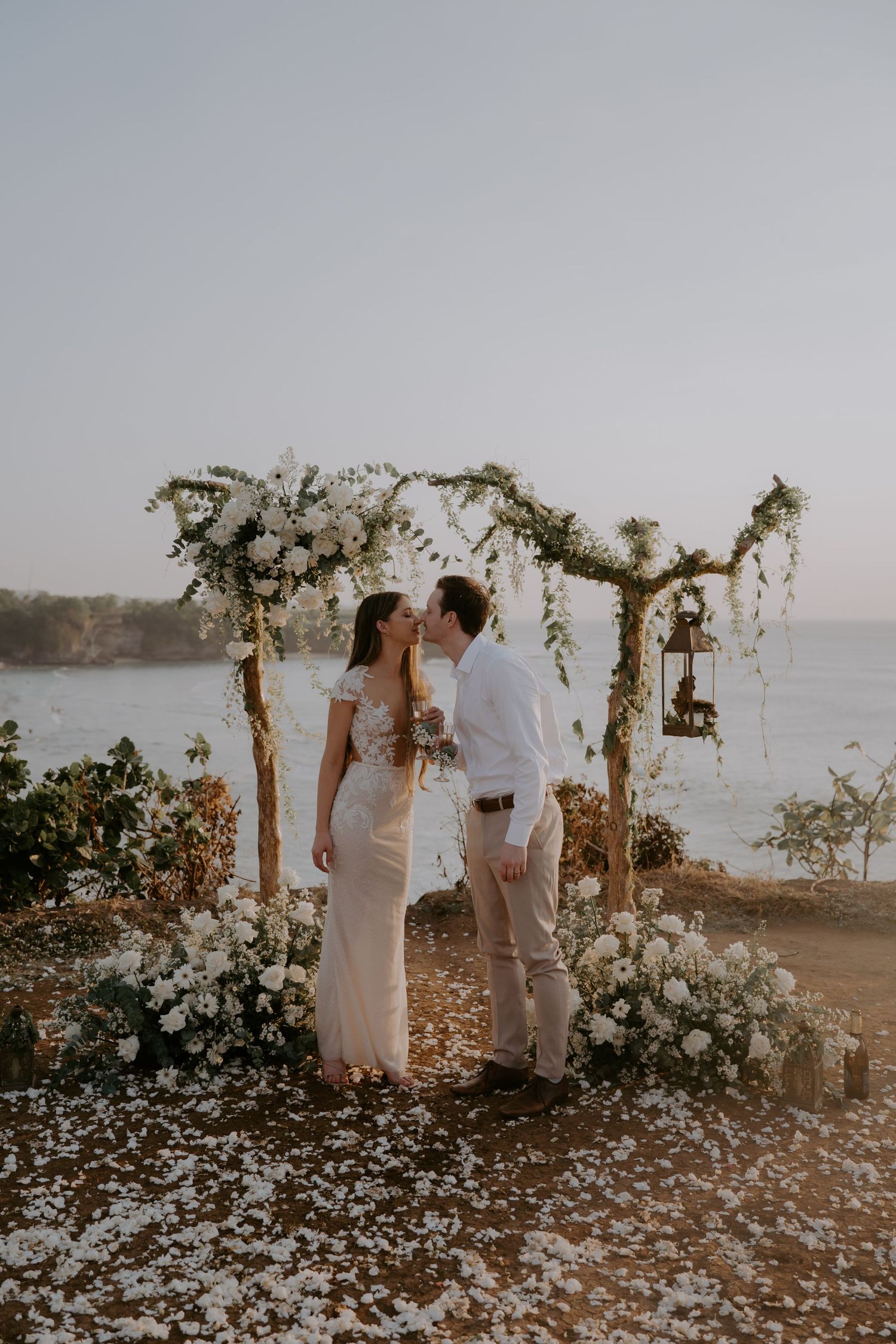 A romantic beach wedding ceremony setup with a floral arch overlooking the ocean at sunset.