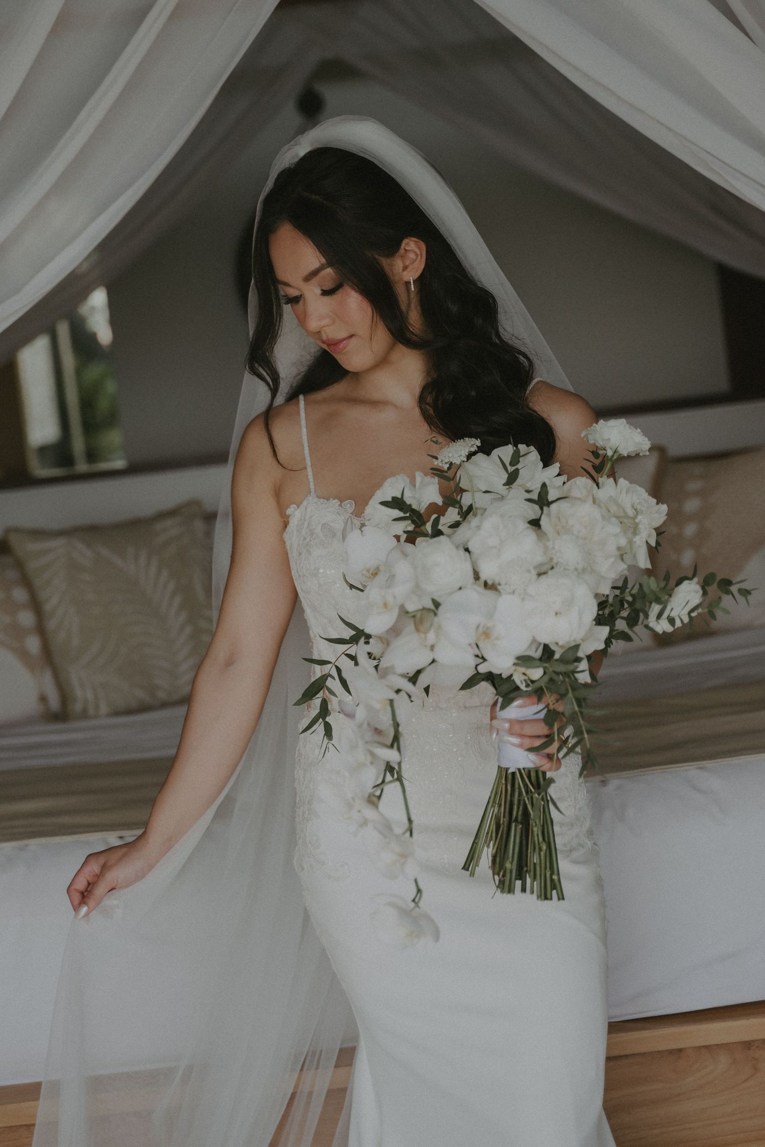 A bride in a fitted white wedding dress holds an elegant bouquet of white orchids and greenery in a sunlit room.