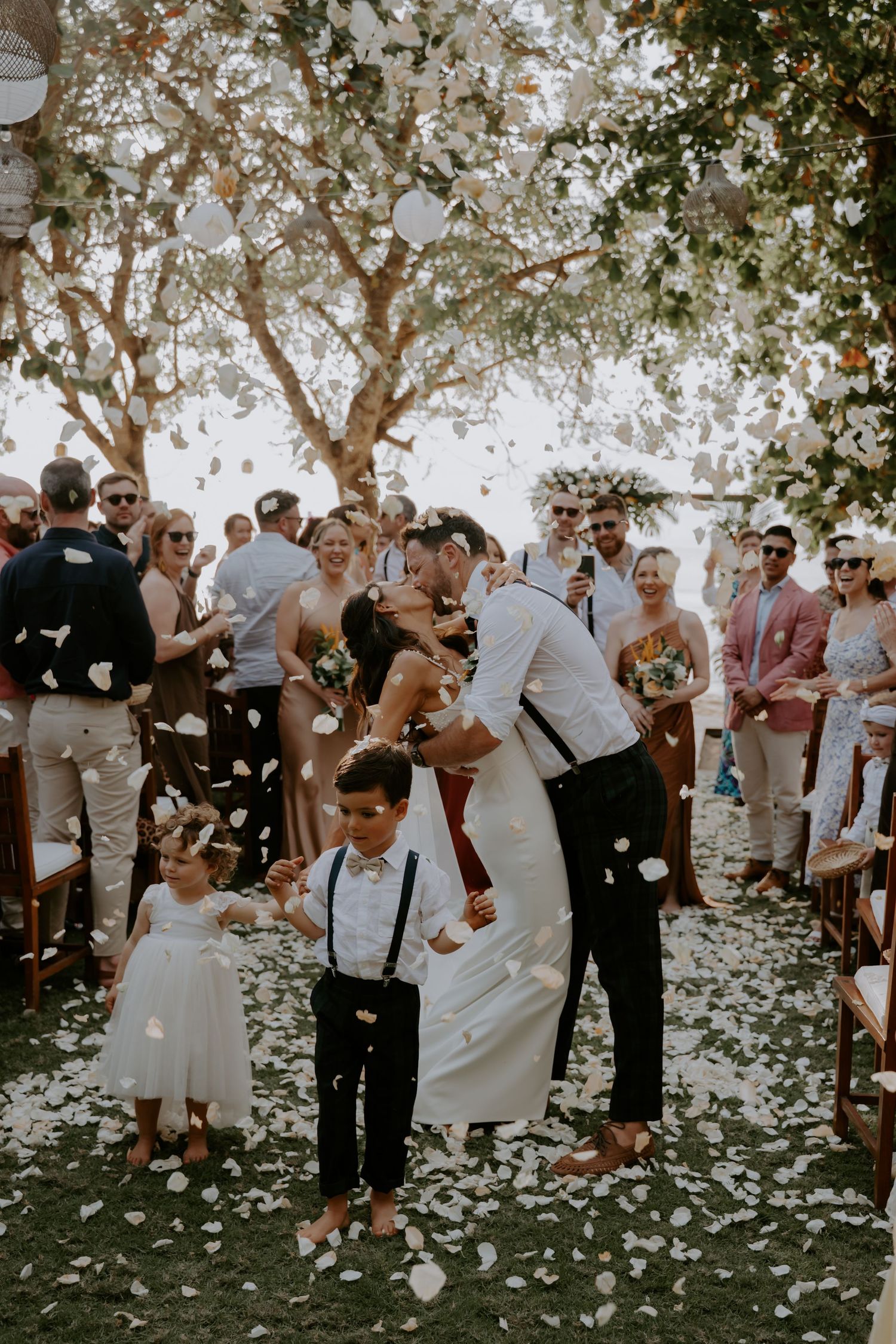 Wedding guests celebrate with white flower petals falling around them during an outdoor garden celebration.