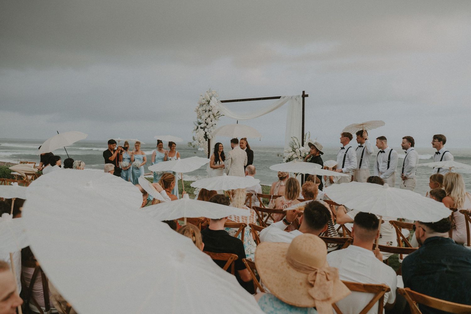 Wedding guests gather on a scenic beach during sunset for an outdoor ceremony with ocean views in the background.