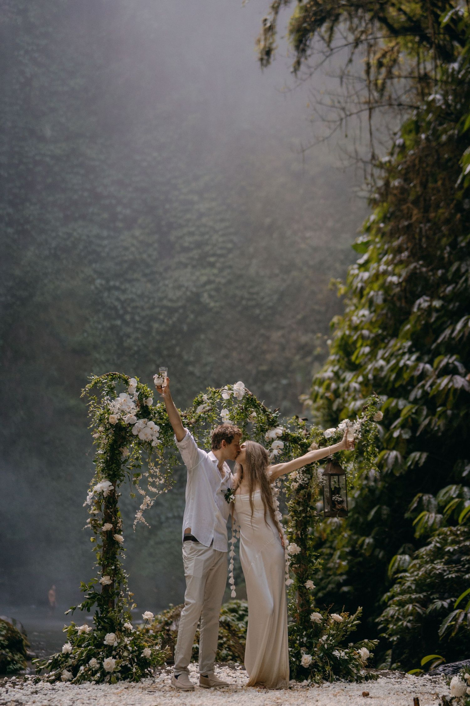 A couple celebrates joyfully with raised arms against a misty forest backdrop adorned with white floral arrangements.