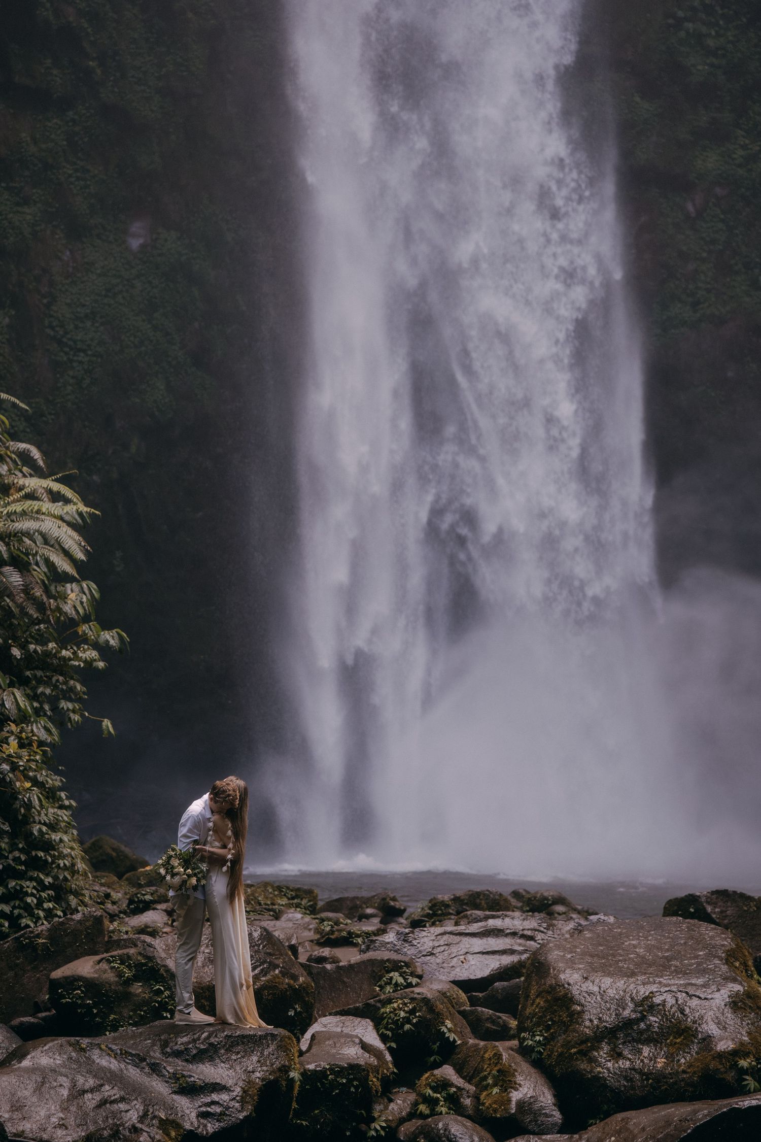 A person in a flowing dress stands near a majestic waterfall on rocky terrain during an atmospheric foggy day.