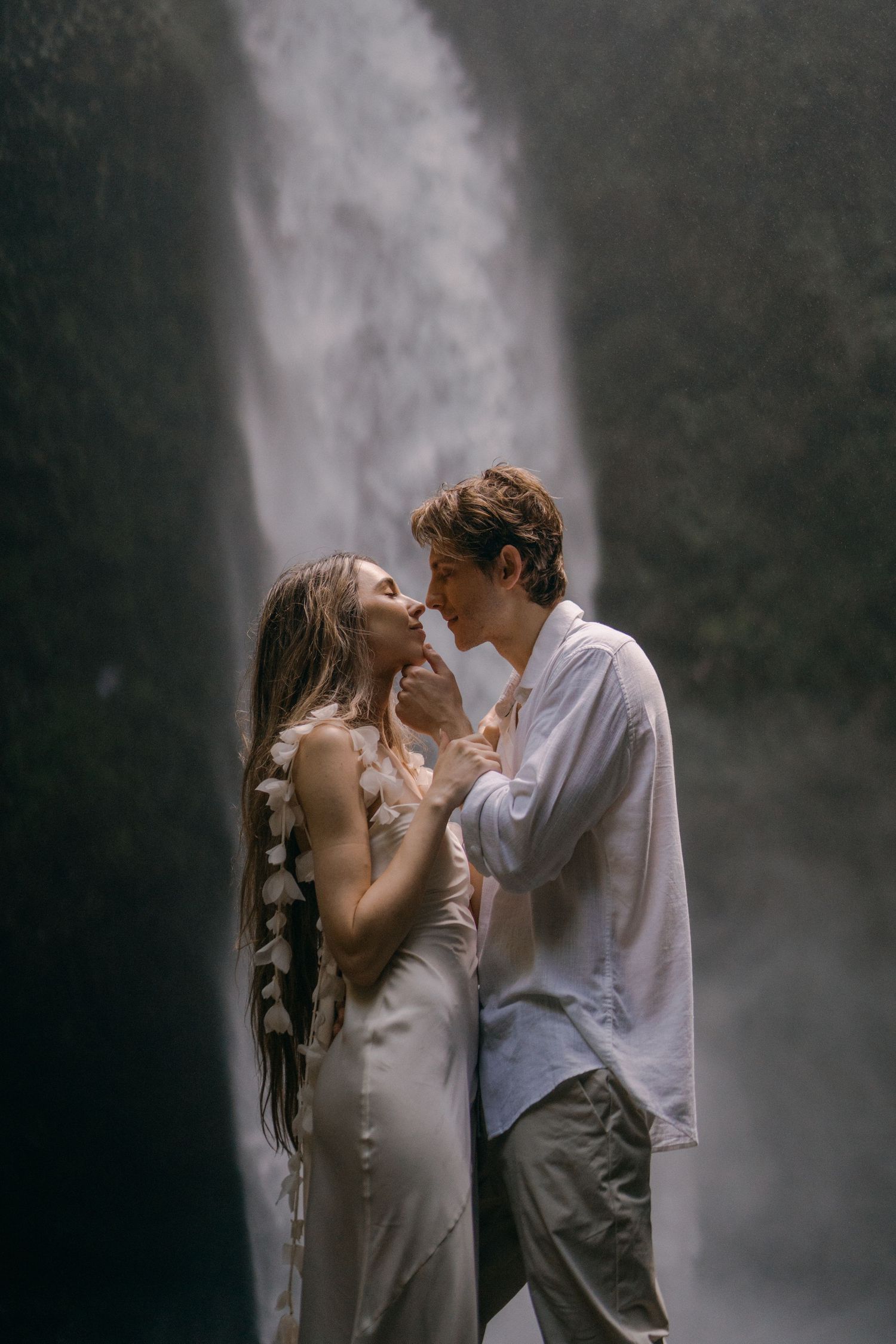 A romantic couple shares an intimate moment in front of a misty waterfall backdrop.