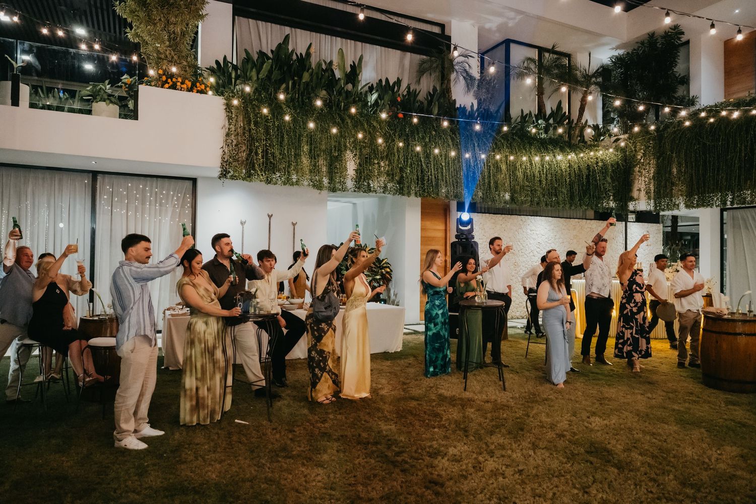Wedding guests raise glasses in a celebratory toast at an elegant indoor reception venue with string lights.