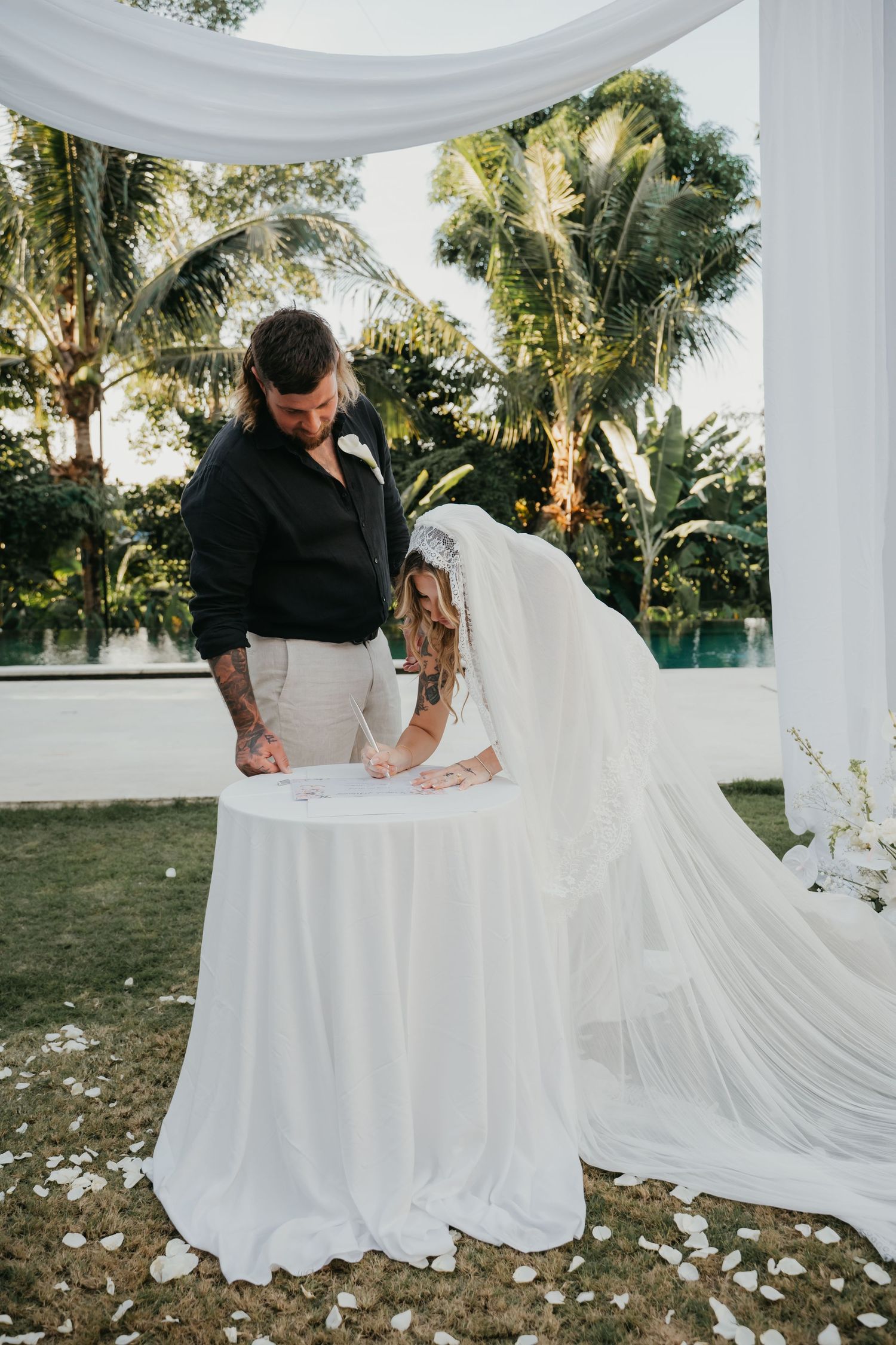 Bride and groom signing wedding registry at a white draped table surrounded by tropical greenery.