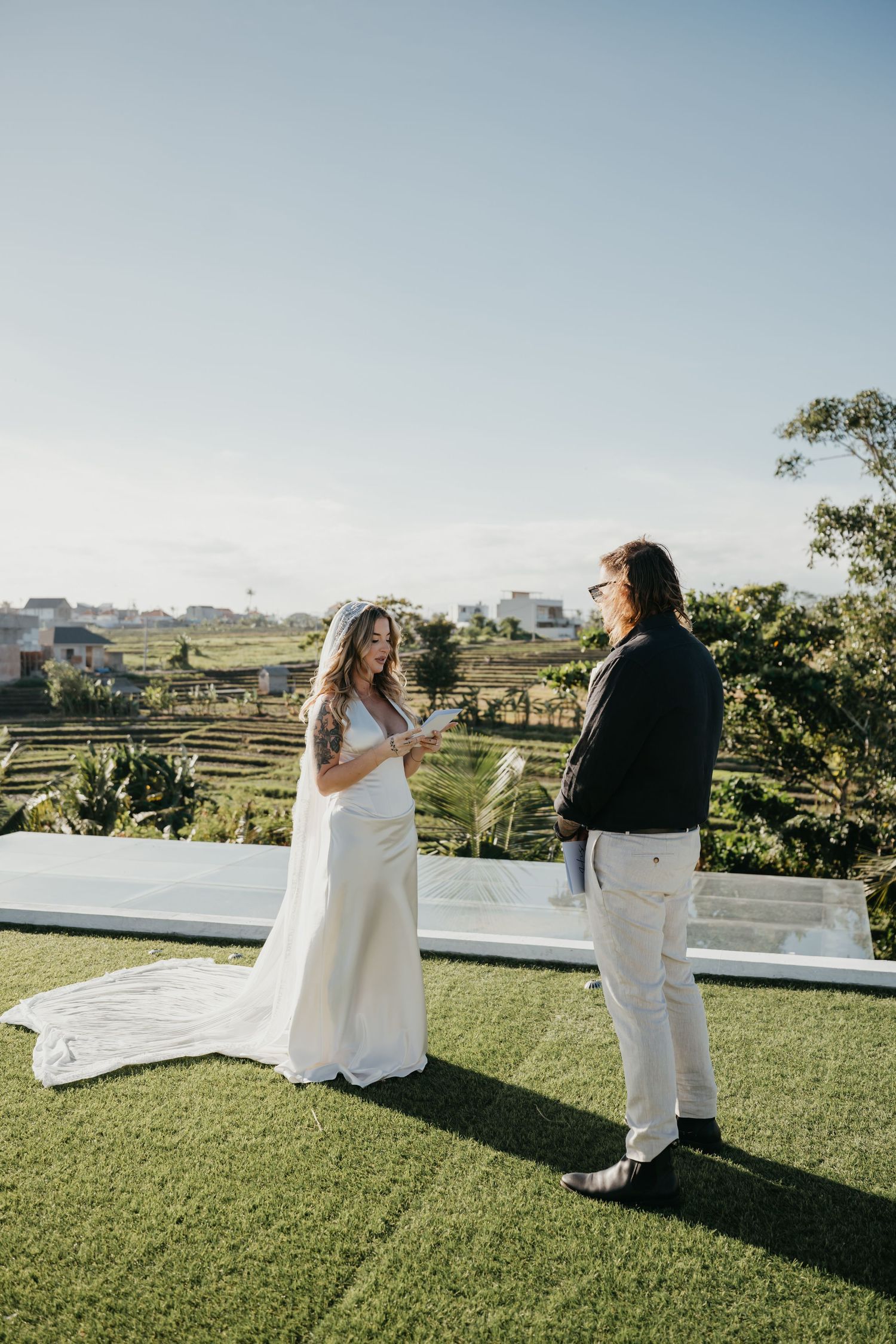Bride reads vows to groom outdoors on green lawn with scenic rice terraces and blue sky backdrop.