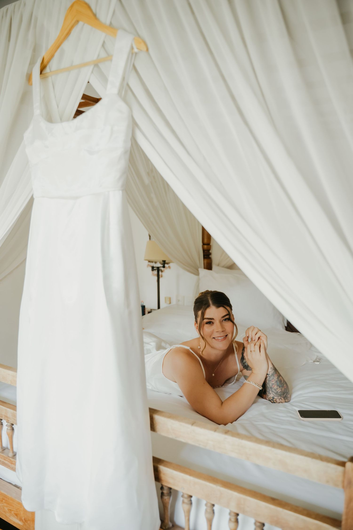 Smiling bride lying on canopy bed beside her elegant white wedding dress on a hanger.