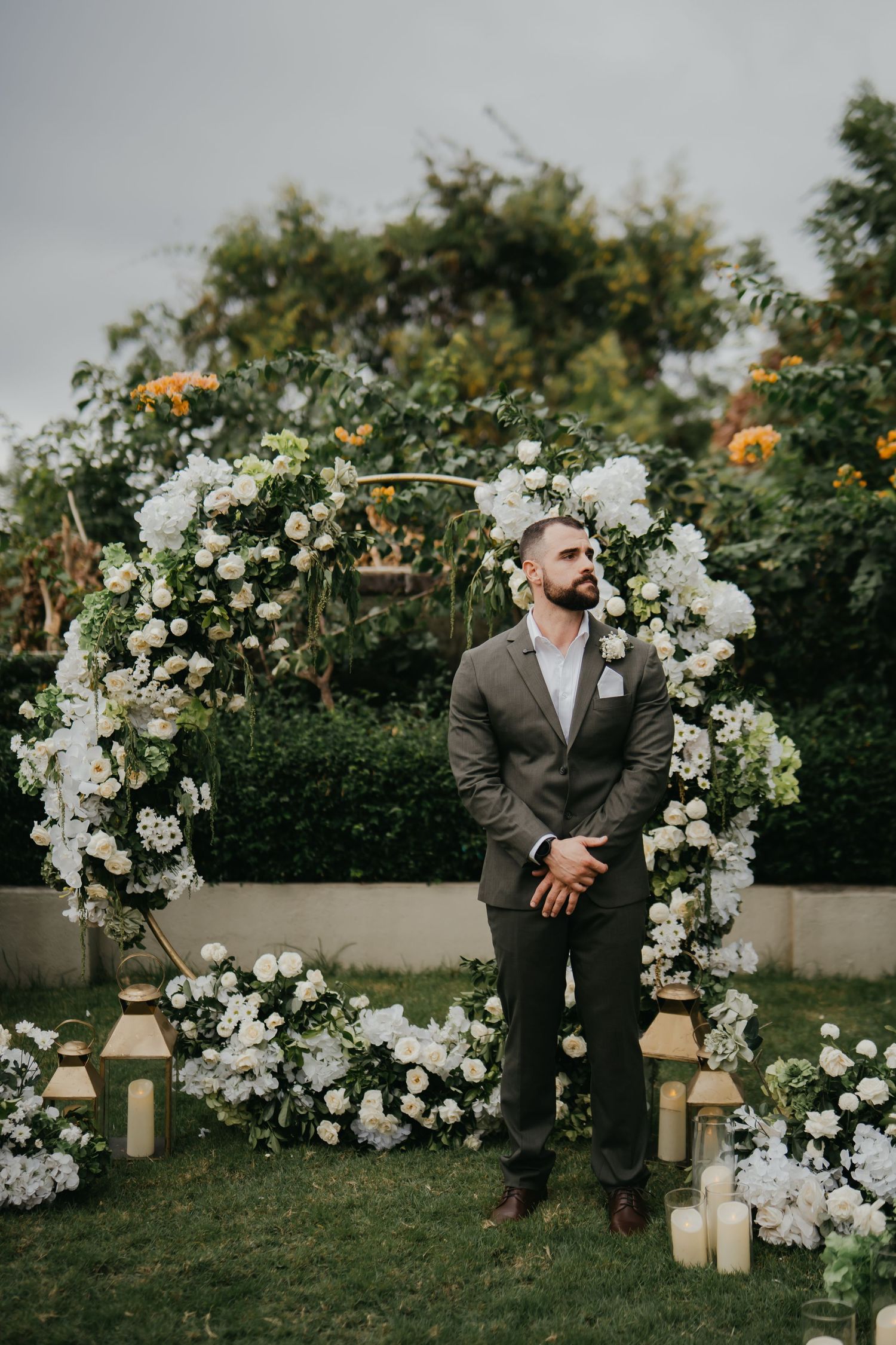 Groom in gray suit stands before circular white floral arch with candles at outdoor ceremony.
