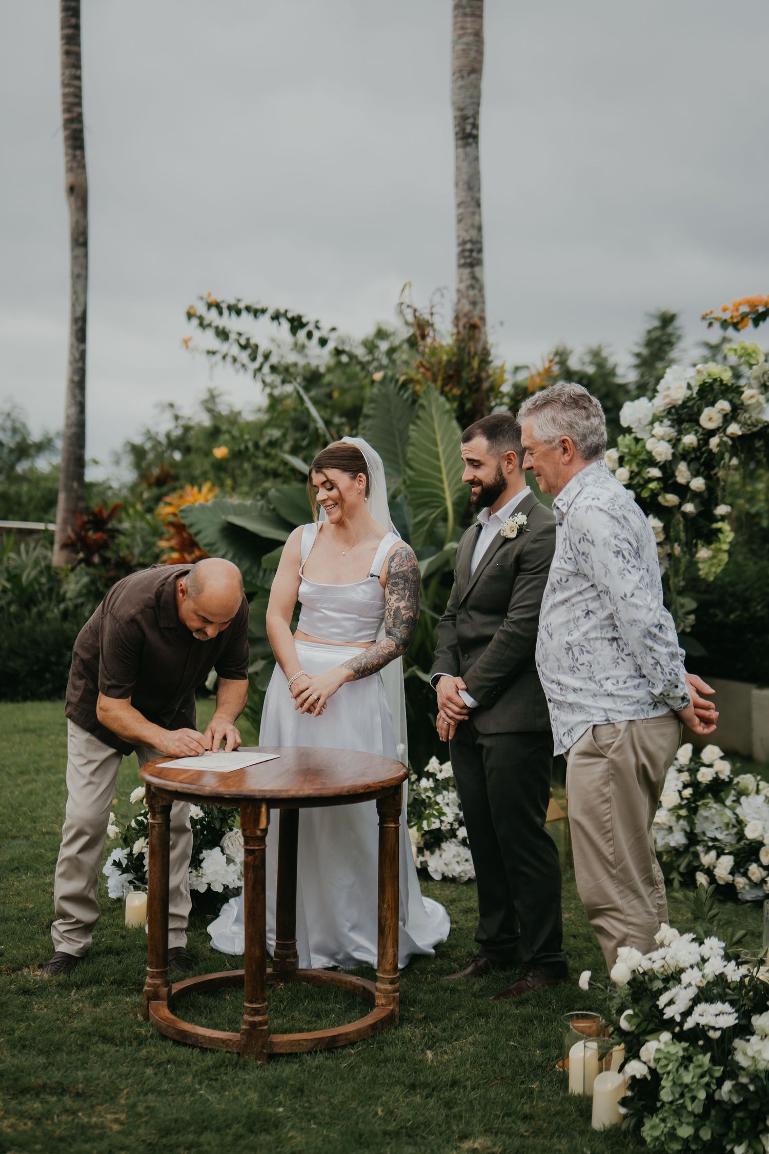 Bride and groom sign wedding register outdoors surrounded by white floral arrangements and palm trees.