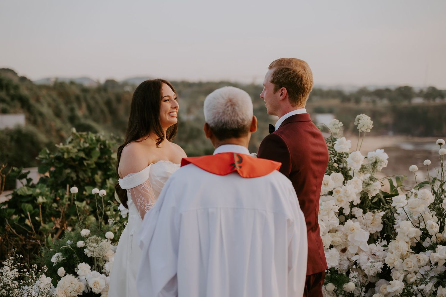 Bride and groom share a joyful moment with officiant during intimate outdoor wedding ceremony at sunset.