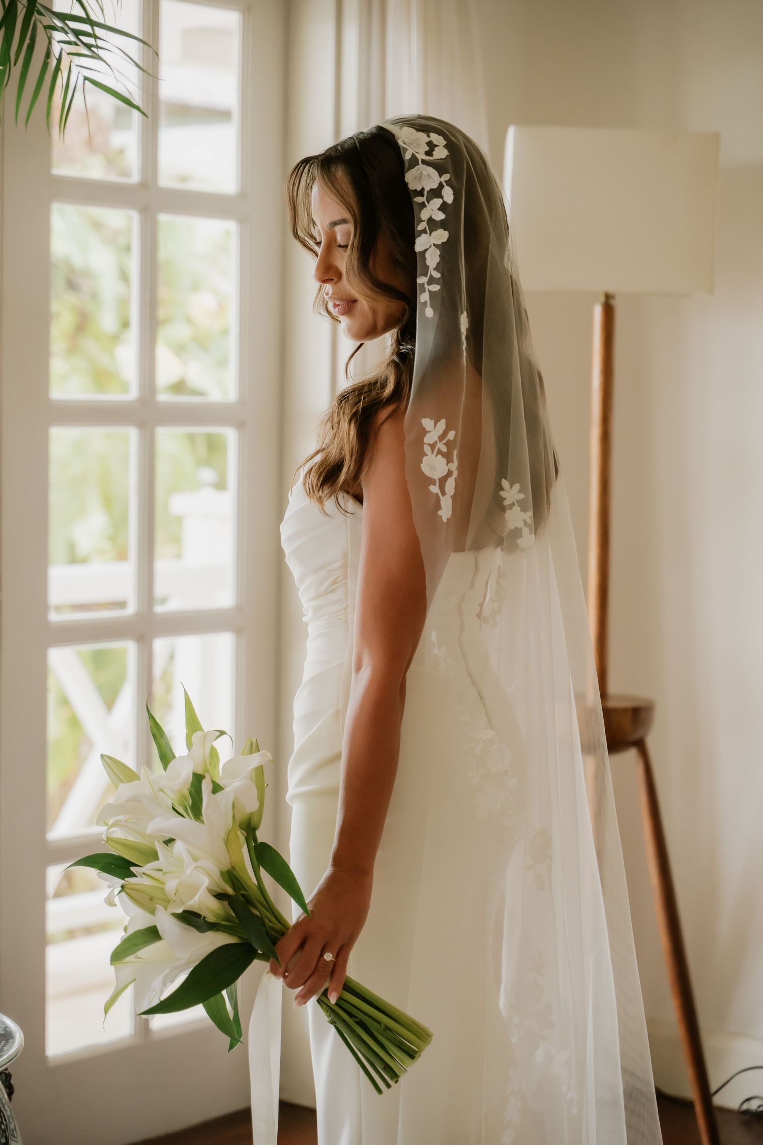 Bride in white strapless gown with lace veil holding white lily bouquet by a sunlit window.