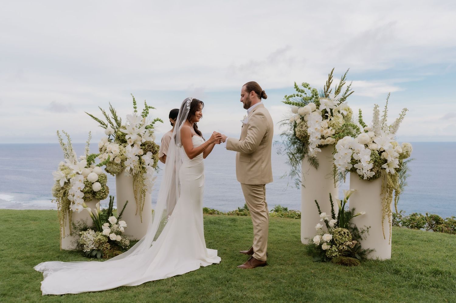 Bride and groom exchange vows at cliffside ceremony with white floral arrangements and ocean backdrop.