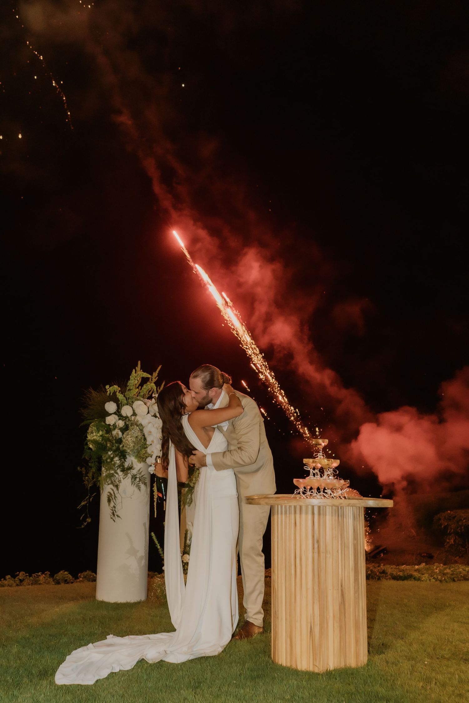 Couple shares romantic kiss beside champagne tower with red fireworks lighting night sky.