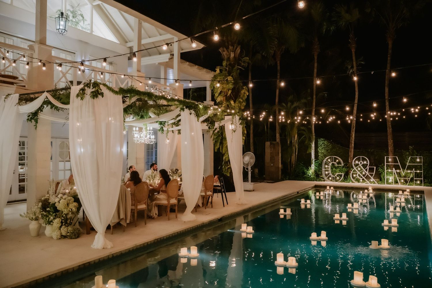 Romantic outdoor wedding reception setup beside a glowing pool with string lights and white drapes.