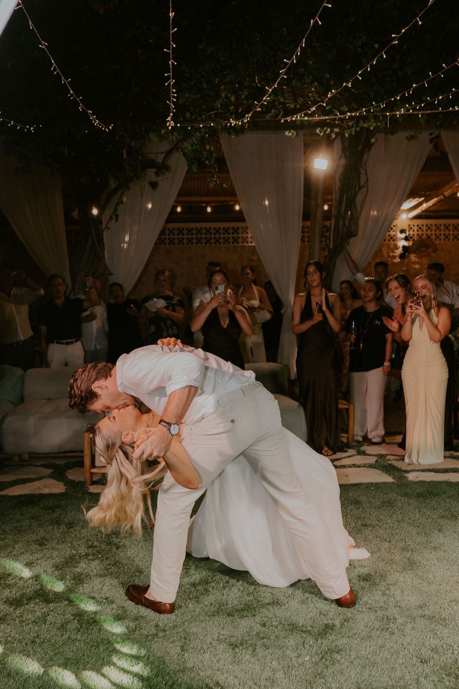 Groom dips bride in romantic first dance kiss surrounded by applauding guests at outdoor wedding reception.