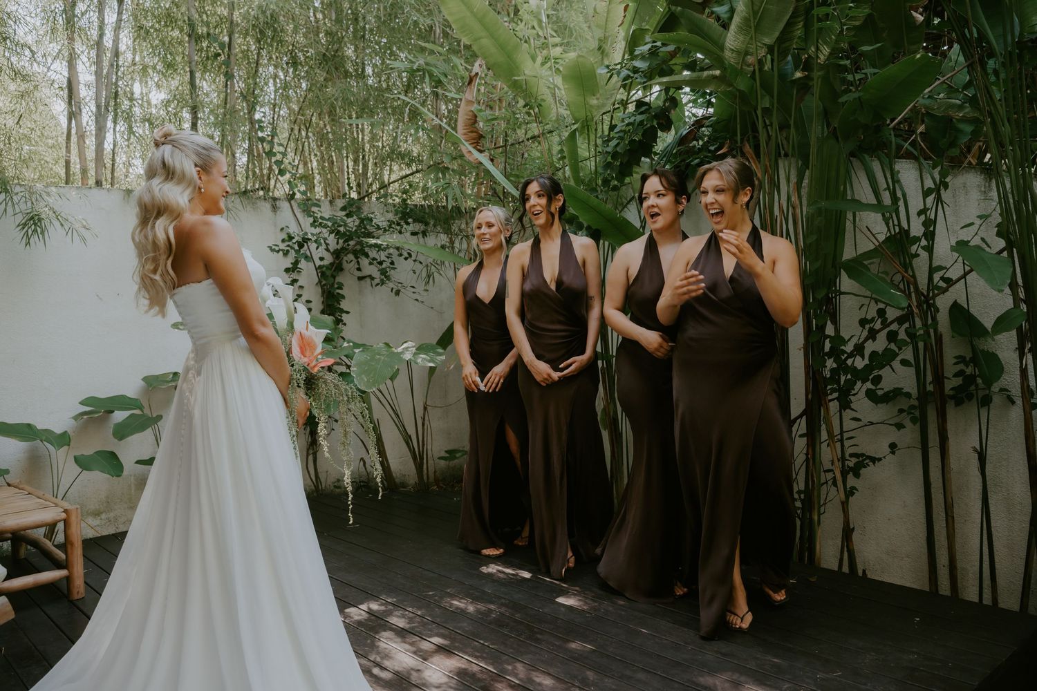 Bridesmaids in brown halter gowns react with joy upon seeing bride in white wedding dress outdoors.