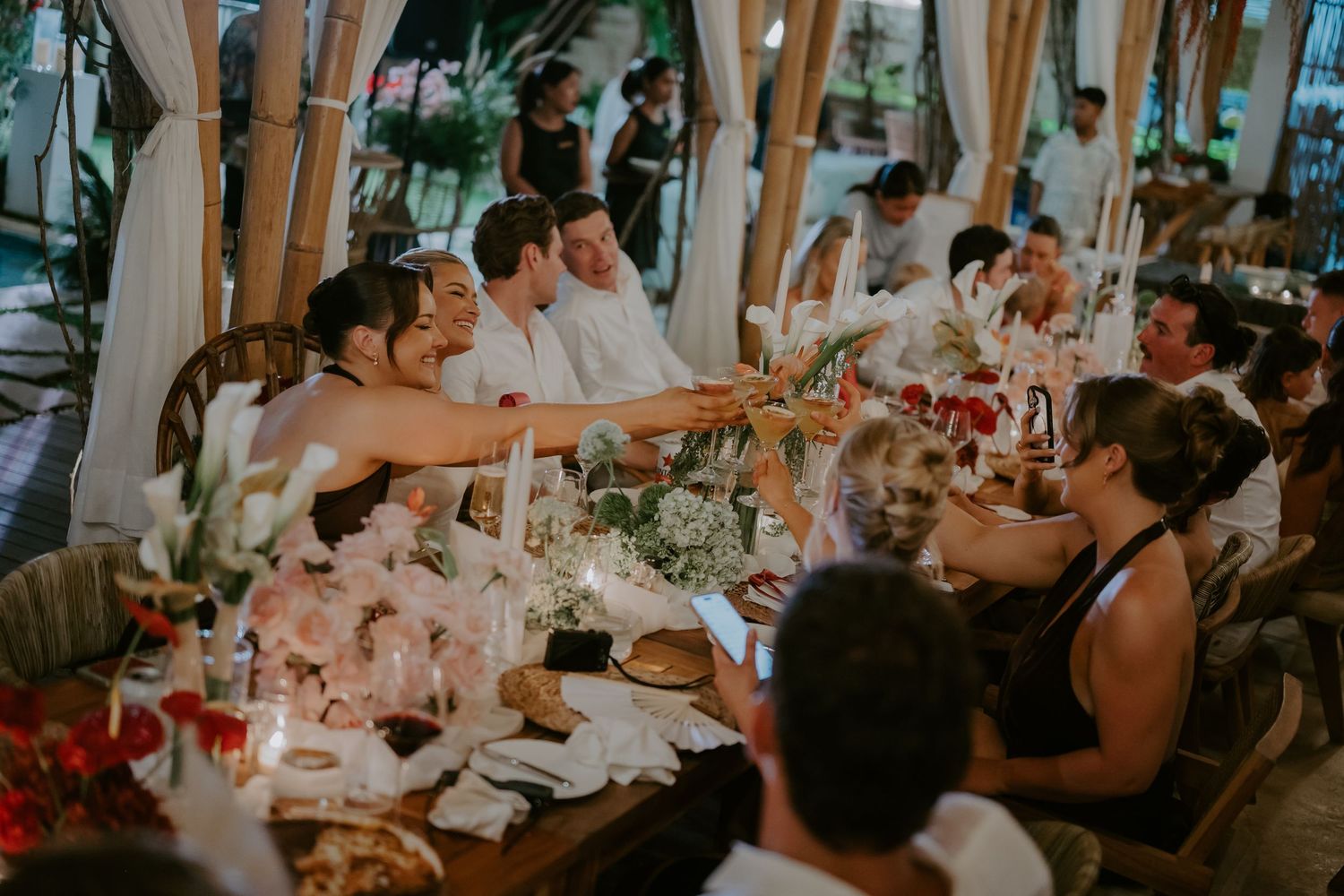 Wedding guests raising glasses in a toast at a candlelit outdoor reception dinner table.