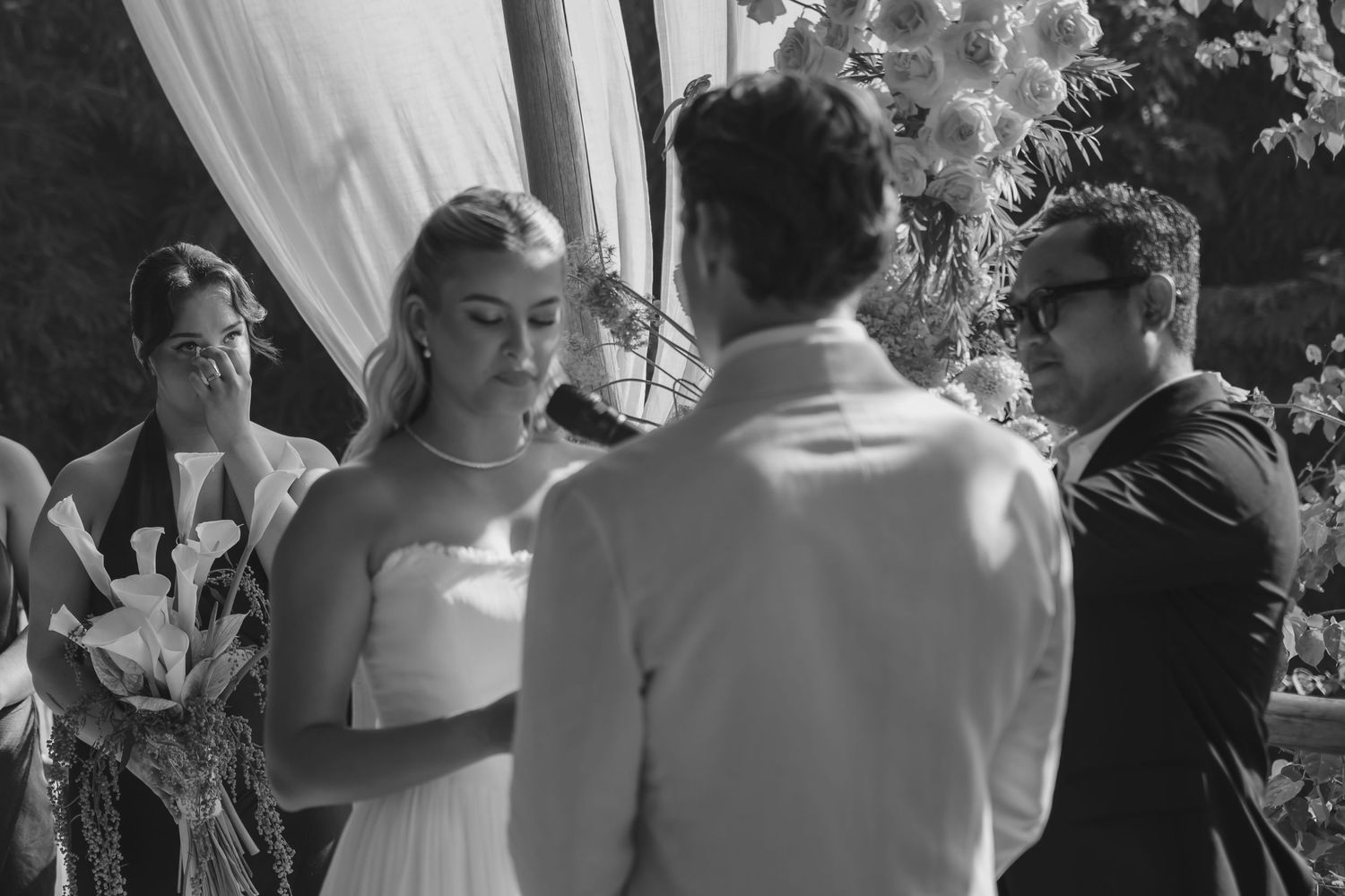 Bride and groom exchange vows at outdoor wedding ceremony with floral arch in black and white photo.