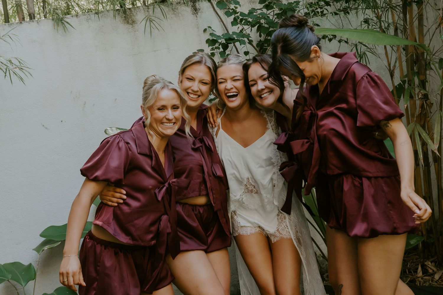 Bride in white lingerie laughing with four bridesmaids wearing matching burgundy satin ruffled sets outdoors.