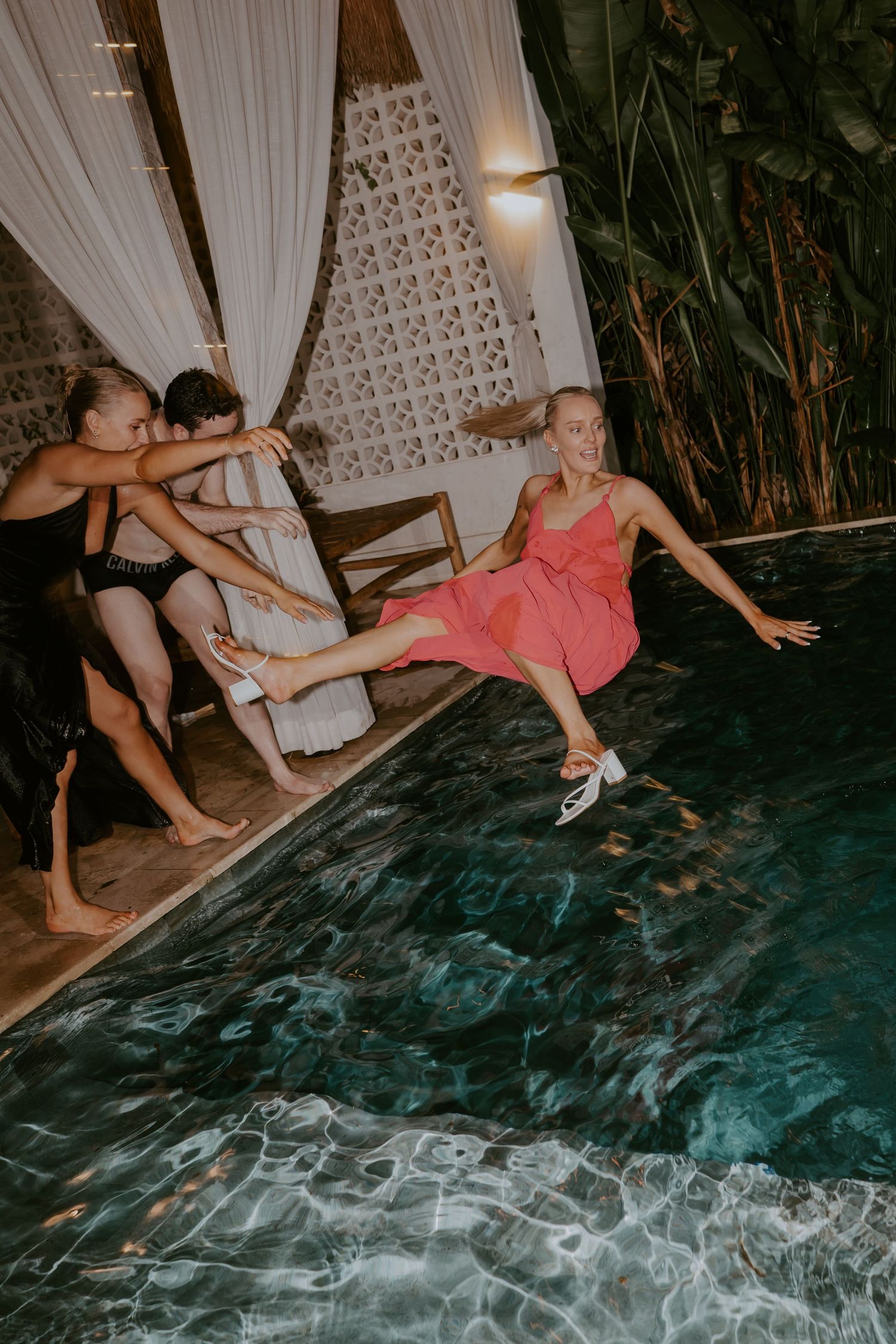 Woman in pink dress being pushed into a pool by friends at a fun wedding after-party.