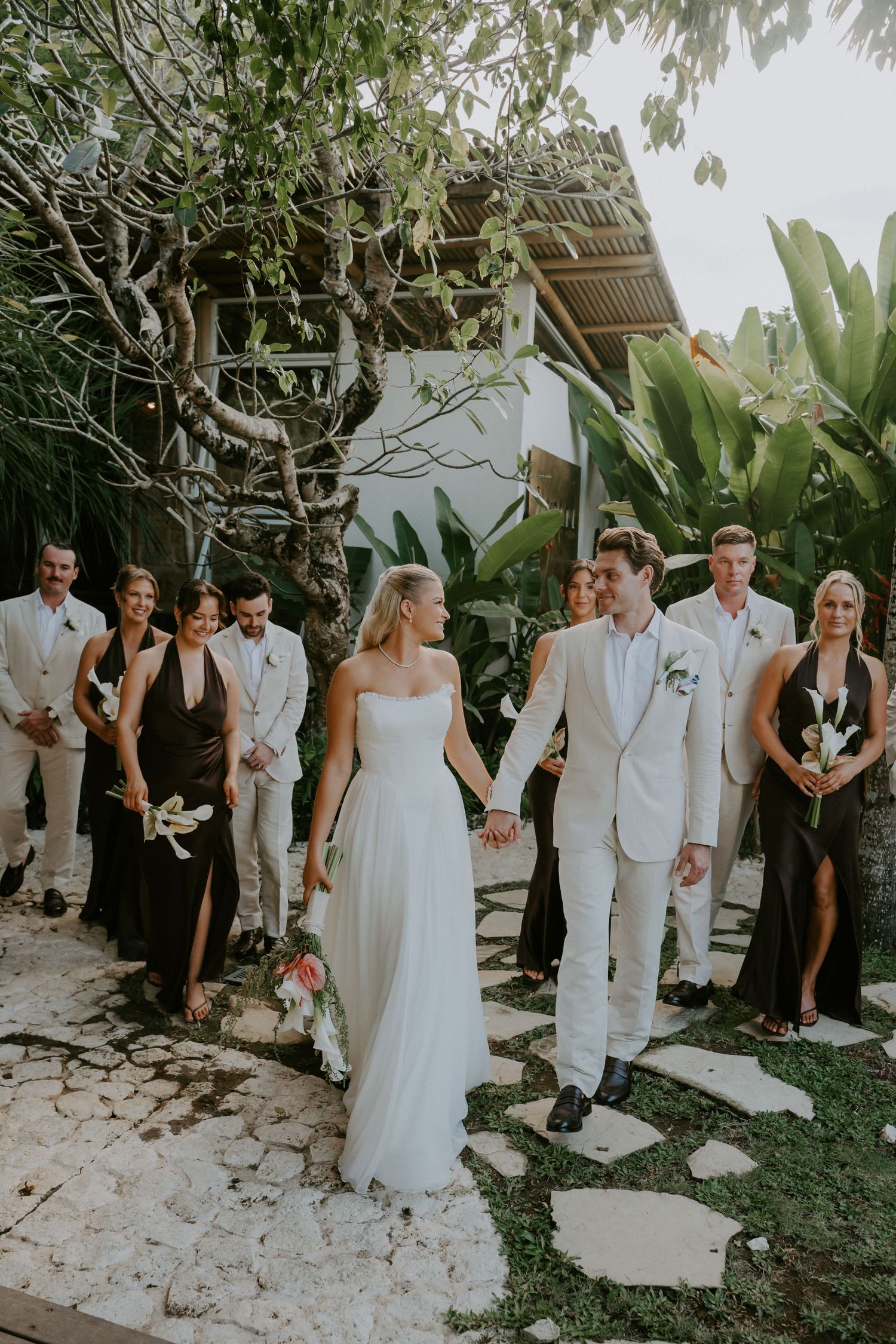 Bride and groom walk hand in hand with wedding party through lush tropical garden venue.