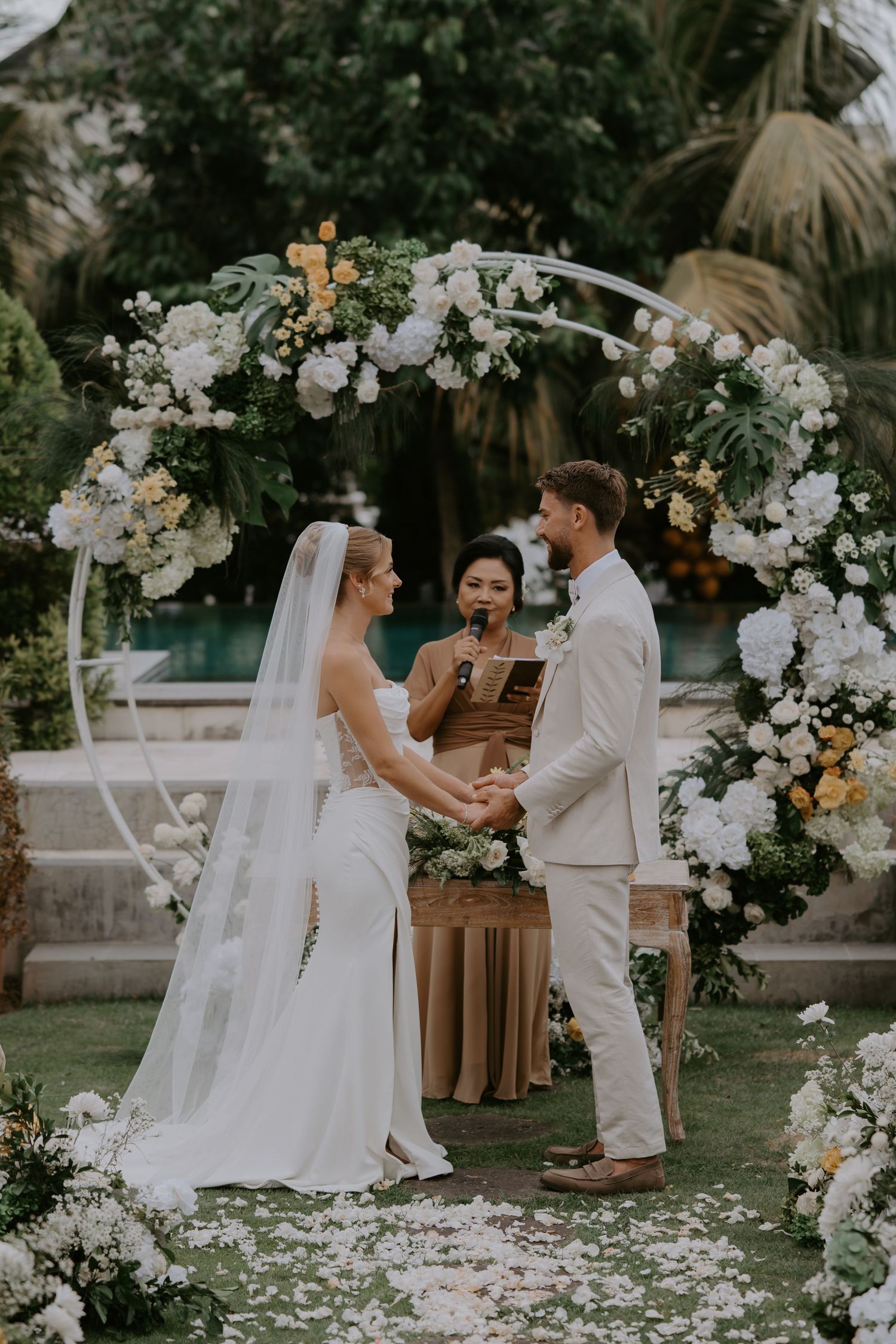 Bride and groom exchange vows under floral arch with white blooms at outdoor garden ceremony.